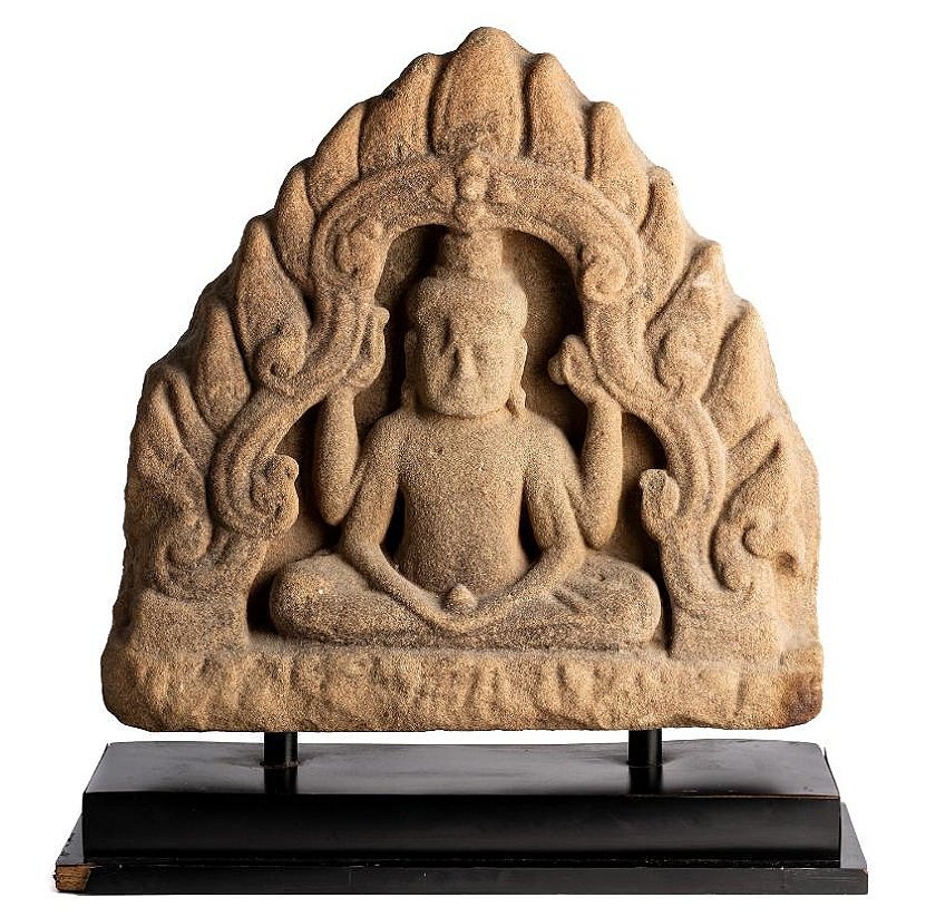 A large grey sandstone relief panel depicting a four-armed deity, probably Avalokiteshvara seated in a sattvasana within a scrolling flaming arch. Cambodia, 12th century C.E.