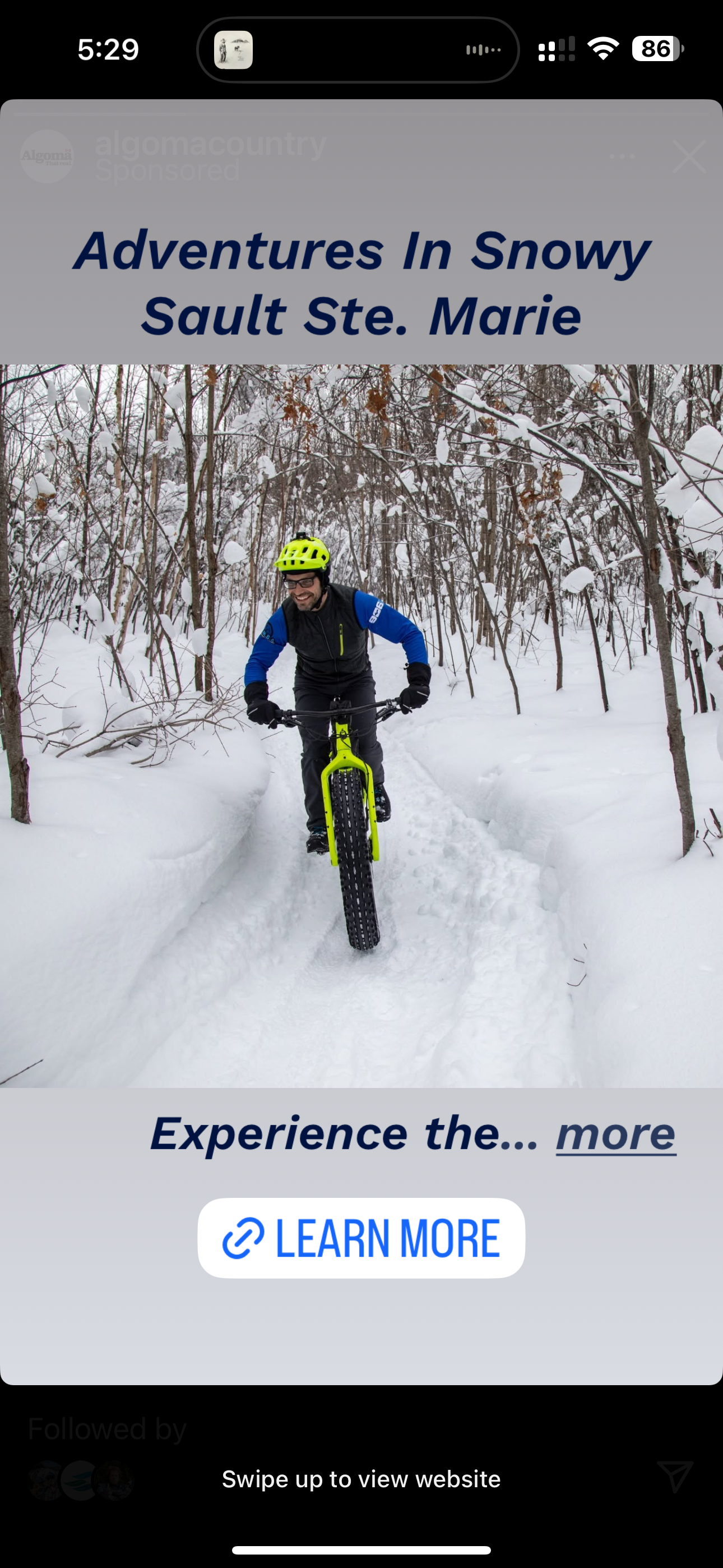 A man riding a fat-tire bicycle on a snow-covered trail in a wooded area during winter. He is wearing a bright yellow helmet, black gloves, and a black jacket with blue sleeves.