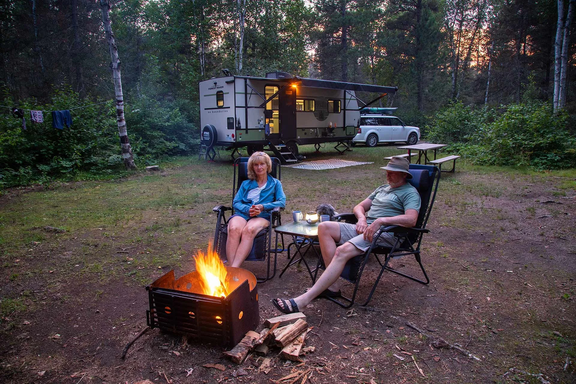 A couple sitting by the campfire with a travel trailer behind them at an Ontario Provincial Park.