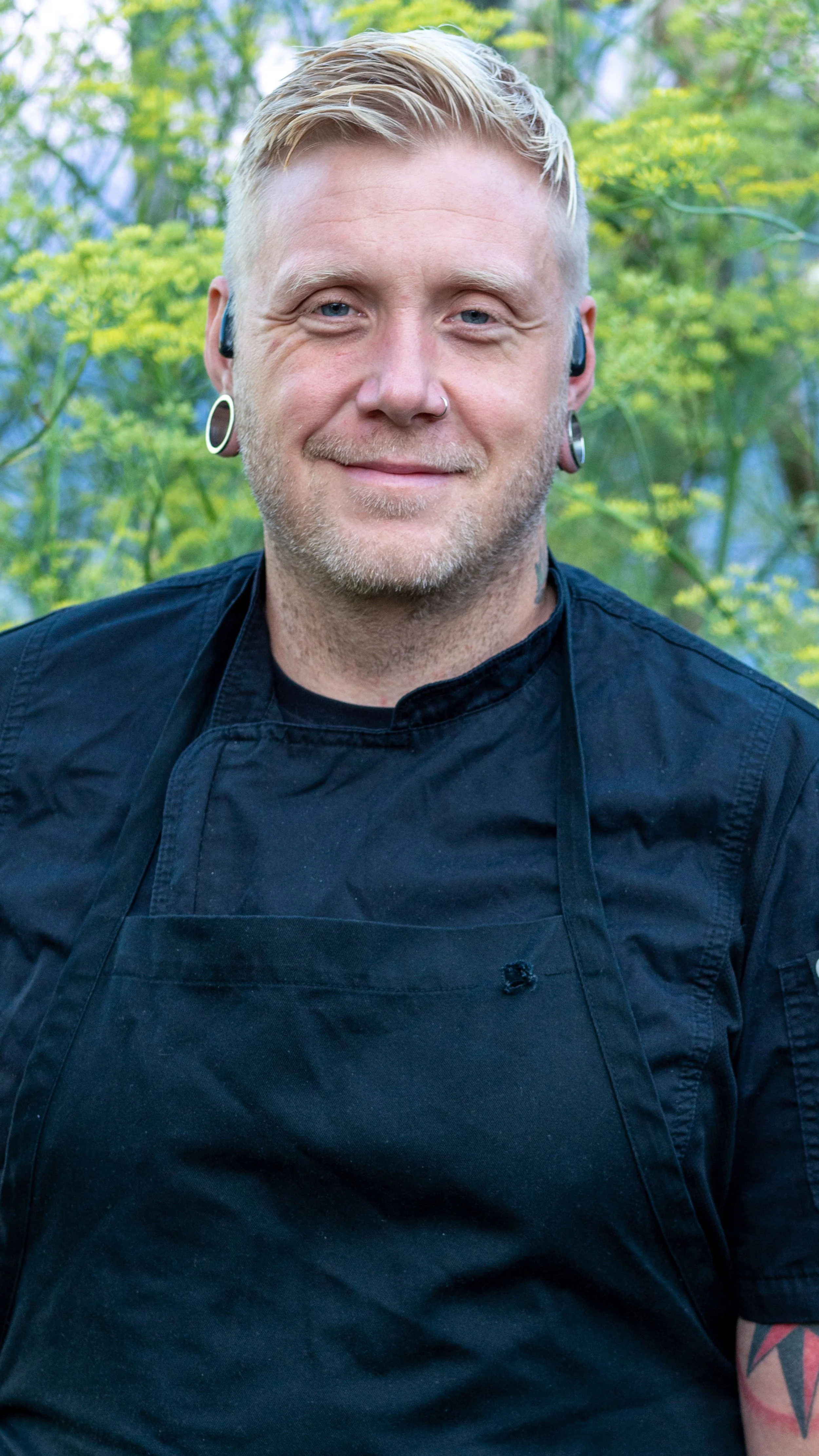 A smiling man wearing a black chef's coat outdoors, with green trees and yellow flowers in the background.