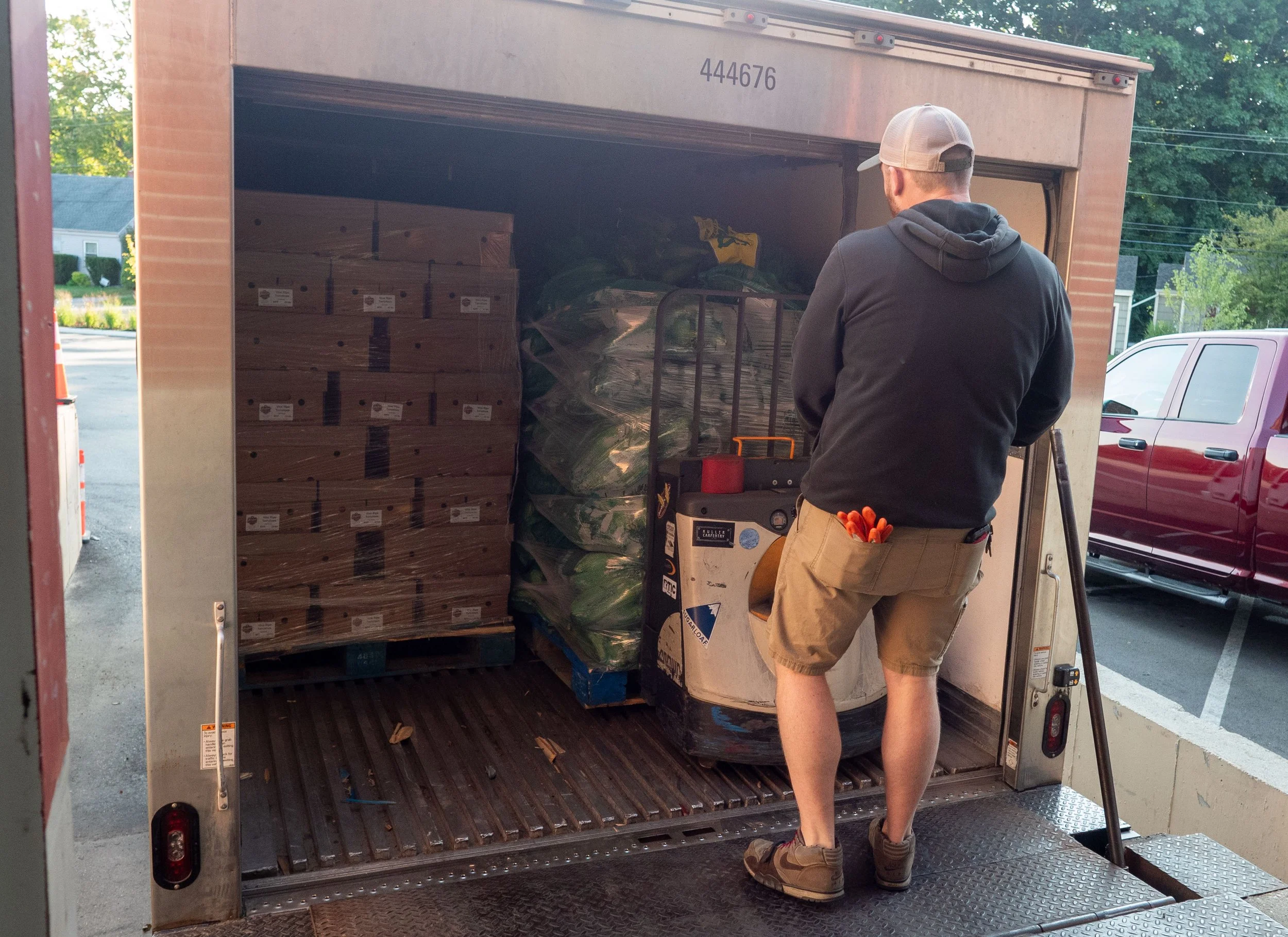 A man with a cap, black hoodie, and beige shorts stands with crossed arms in front of an open moving truck, which is filled with boxes and bags, near parked cars and a suburban street.