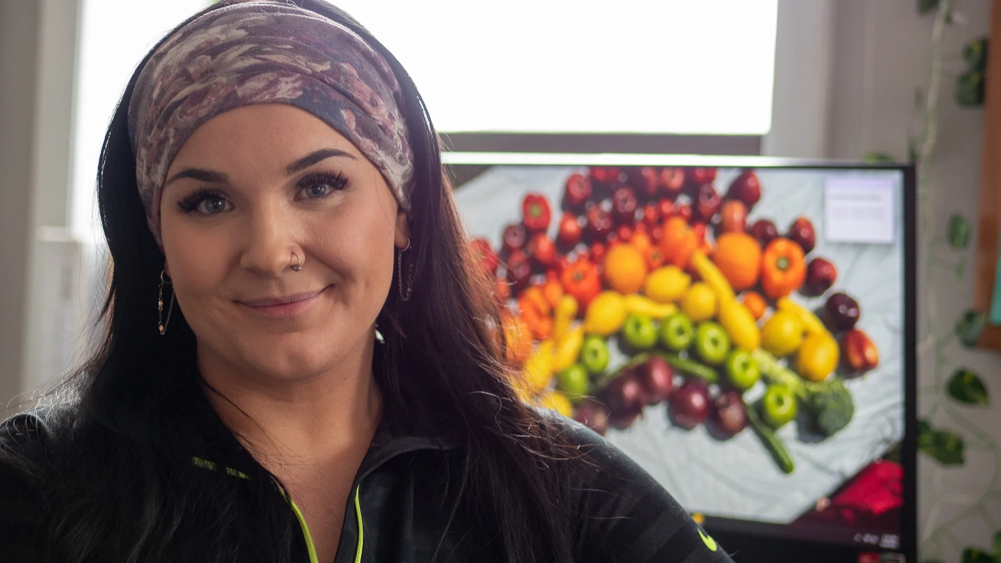 A woman with long dark hair, wearing earrings and a floral headscarf, smiling in front of a screen displaying a colorful arrangement of fruits including red grapes, oranges, yellow bananas, and green kiwis.