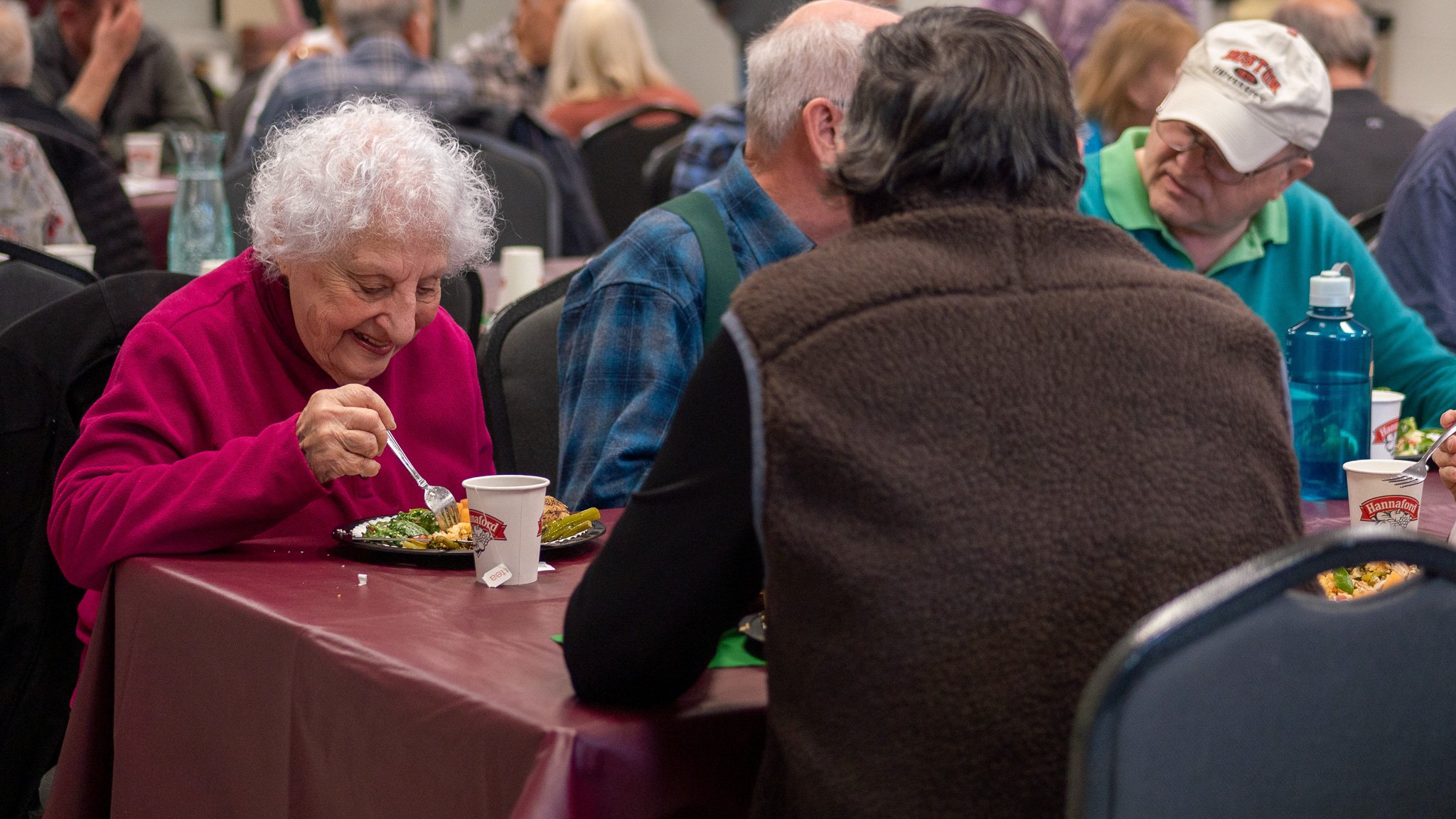 An elderly woman wearing a bright pink sweater is sitting at a table, enjoying a meal with a smile. She has white, curly hair and is using a fork to eat from a black plate with various foods. Several other people are sitting nearby at the same table,