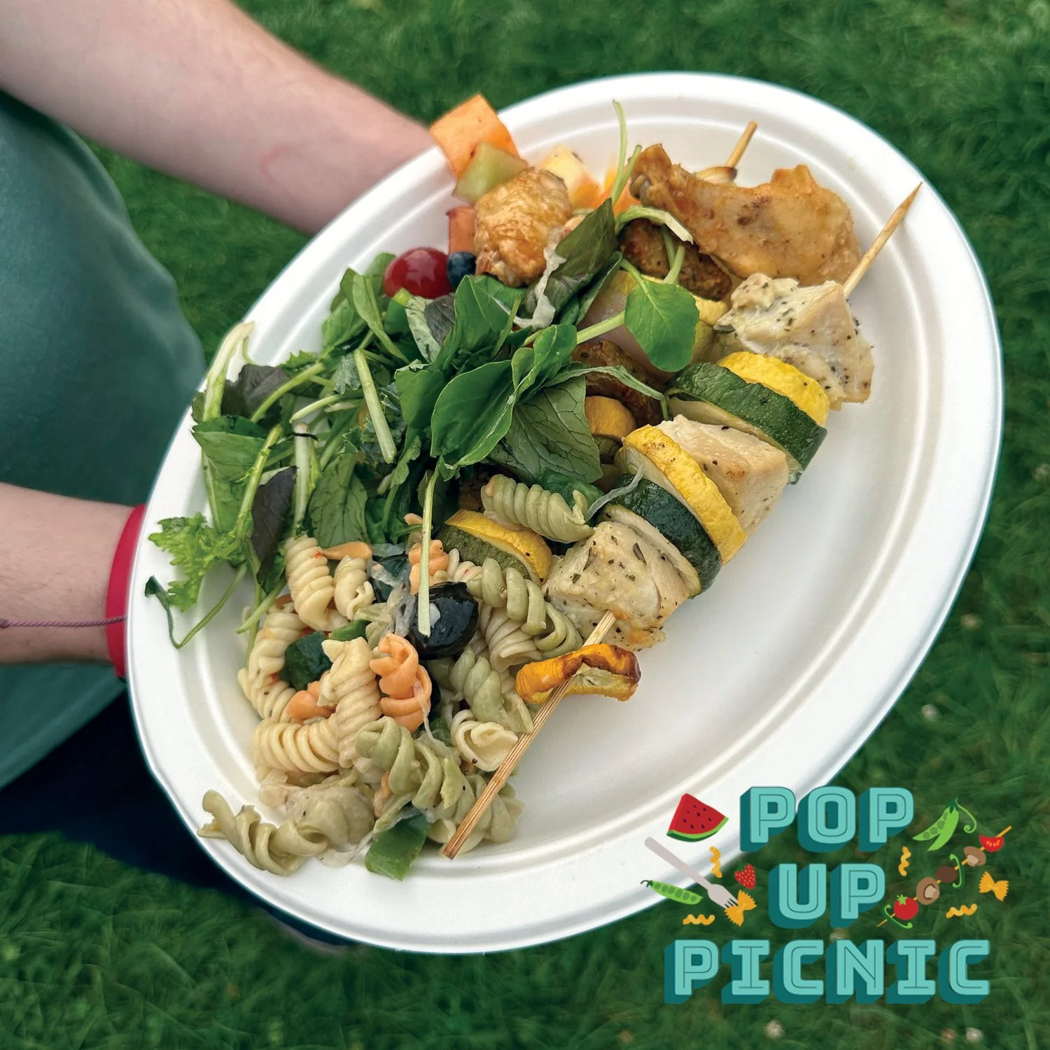 A white disposable plate with grilled chicken skewers, a green leafy salad, and colorful pasta salad, held outdoors on grass with a 'Pop Up Picnic' graphic overlay.