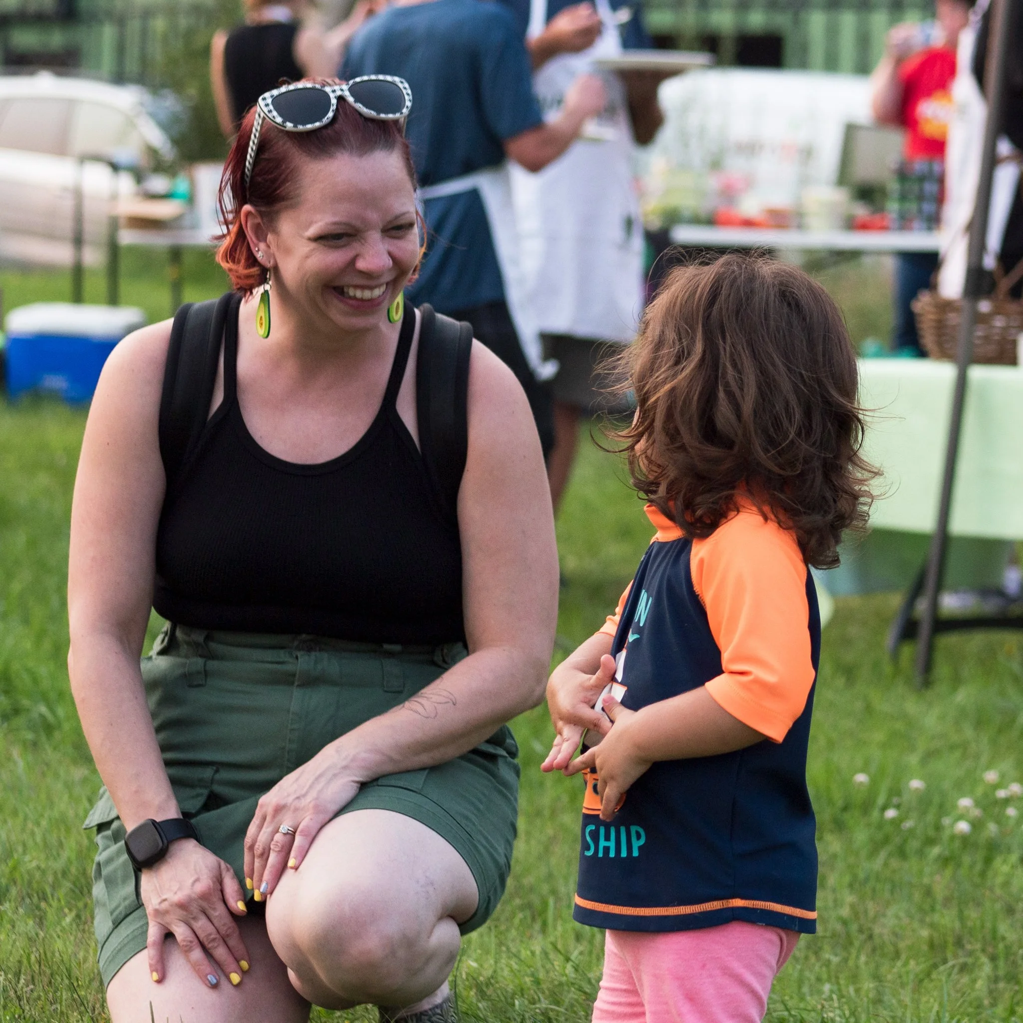 A woman with short red hair, sunglasses on her head, and a black tank top, smiling and talking to a young girl with curly brown hair, wearing a navy and orange shirt and pink shorts, in an outdoor setting with trees and tents.