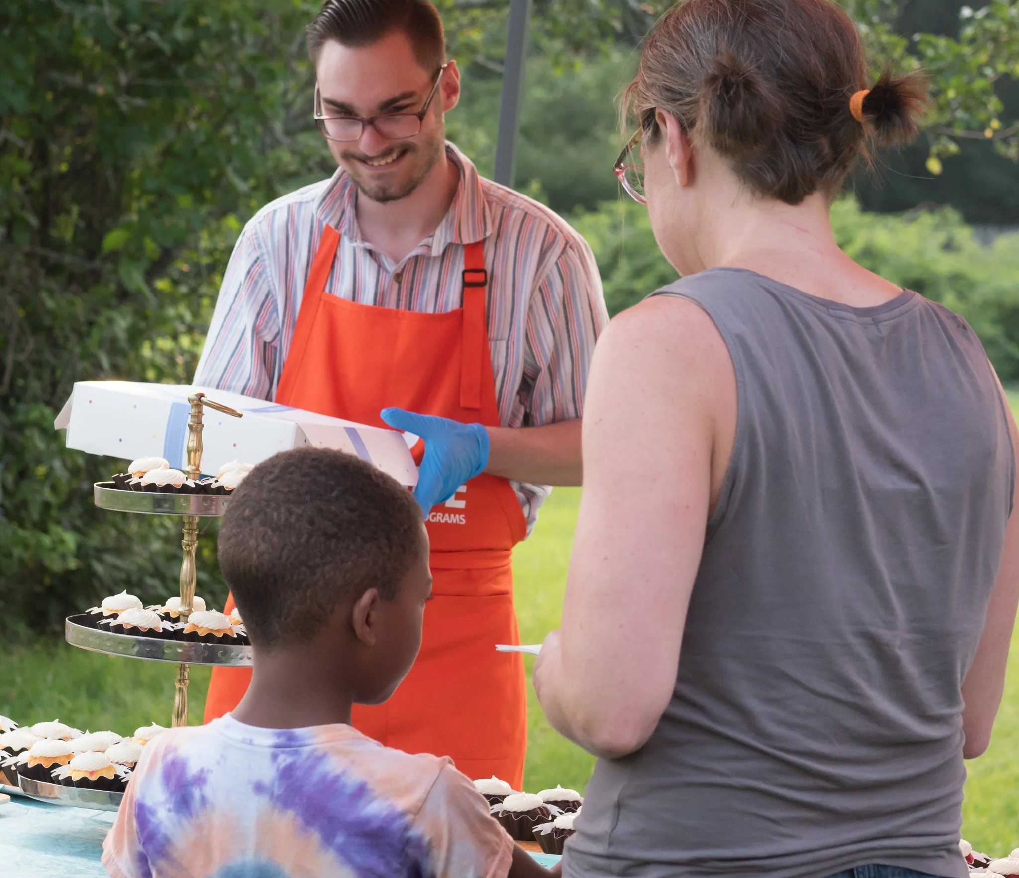 A young man in an orange apron and glasses giving cupcakes to a woman at an outdoor gathering.