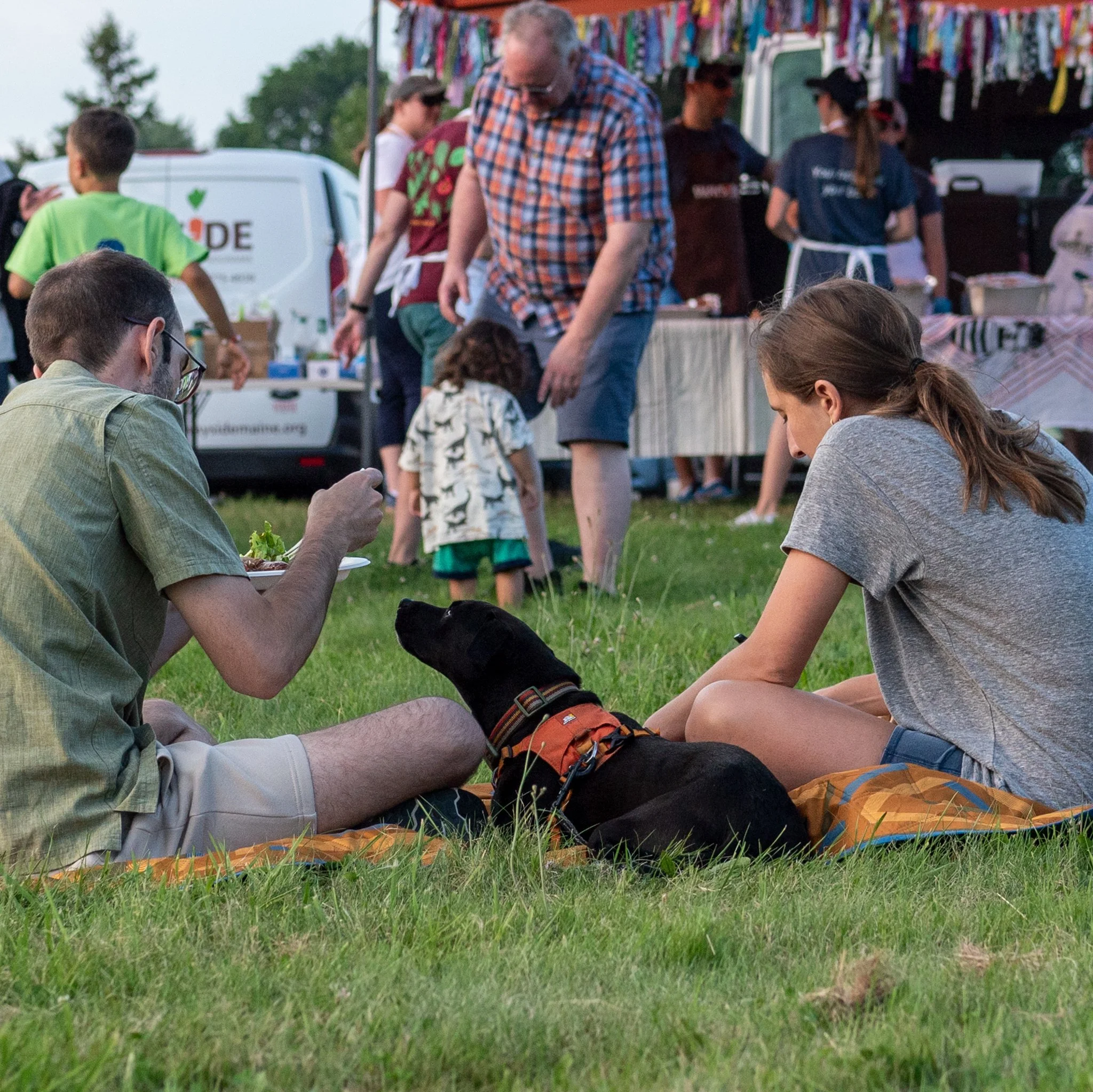 People sitting on the grass at an outdoor event, with a black dog lying between them, surrounded by food and a busy fair or festival scene in the background.