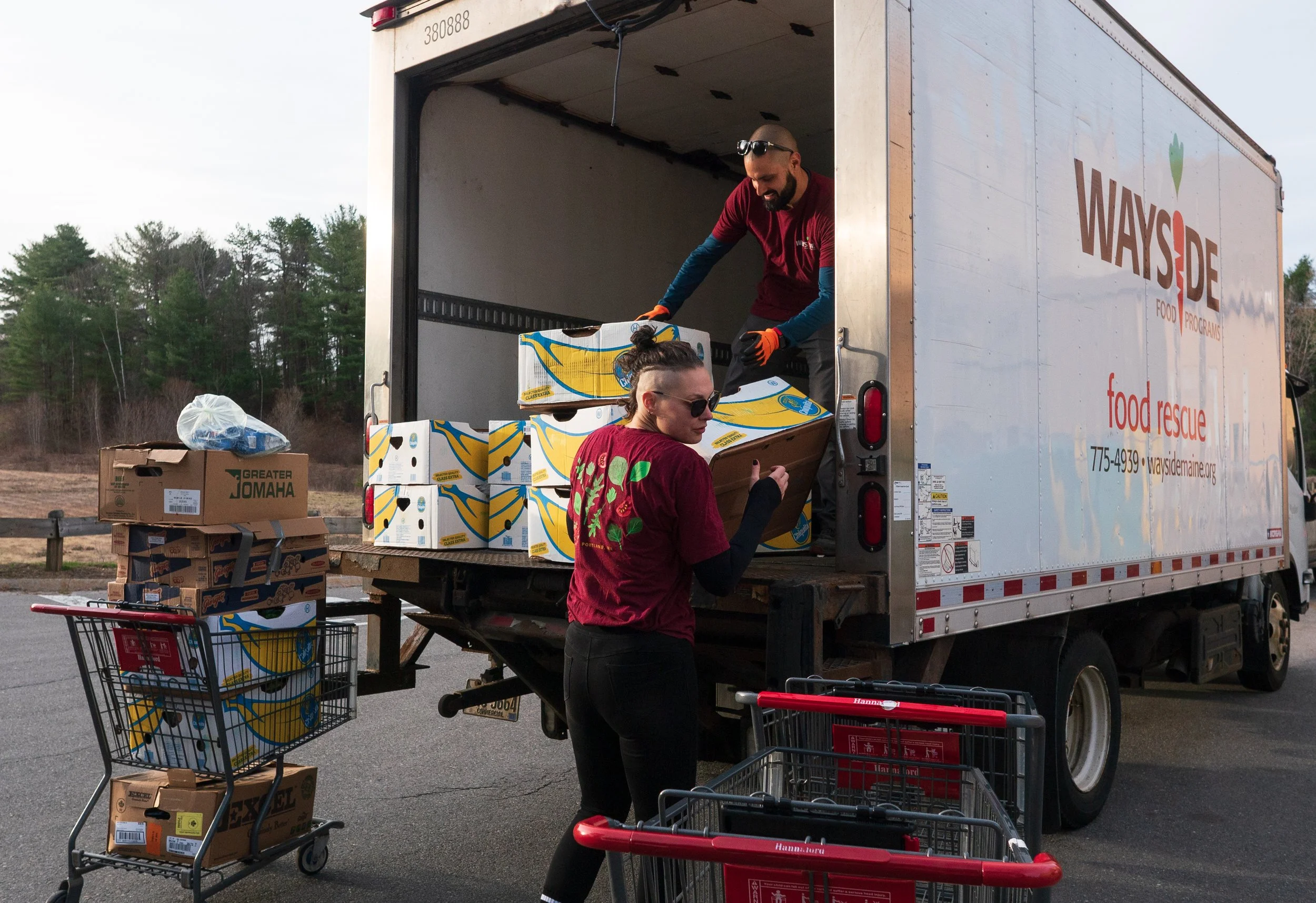 Two volunteers loading boxes of food into a truck marked "Wayside Food Rescue". One person is inside the truck handing down a box to another person standing below, who is holding a box in her hands. There are several grocery carts filled with boxes and bags nearby. The background shows trees and an overcast sky.