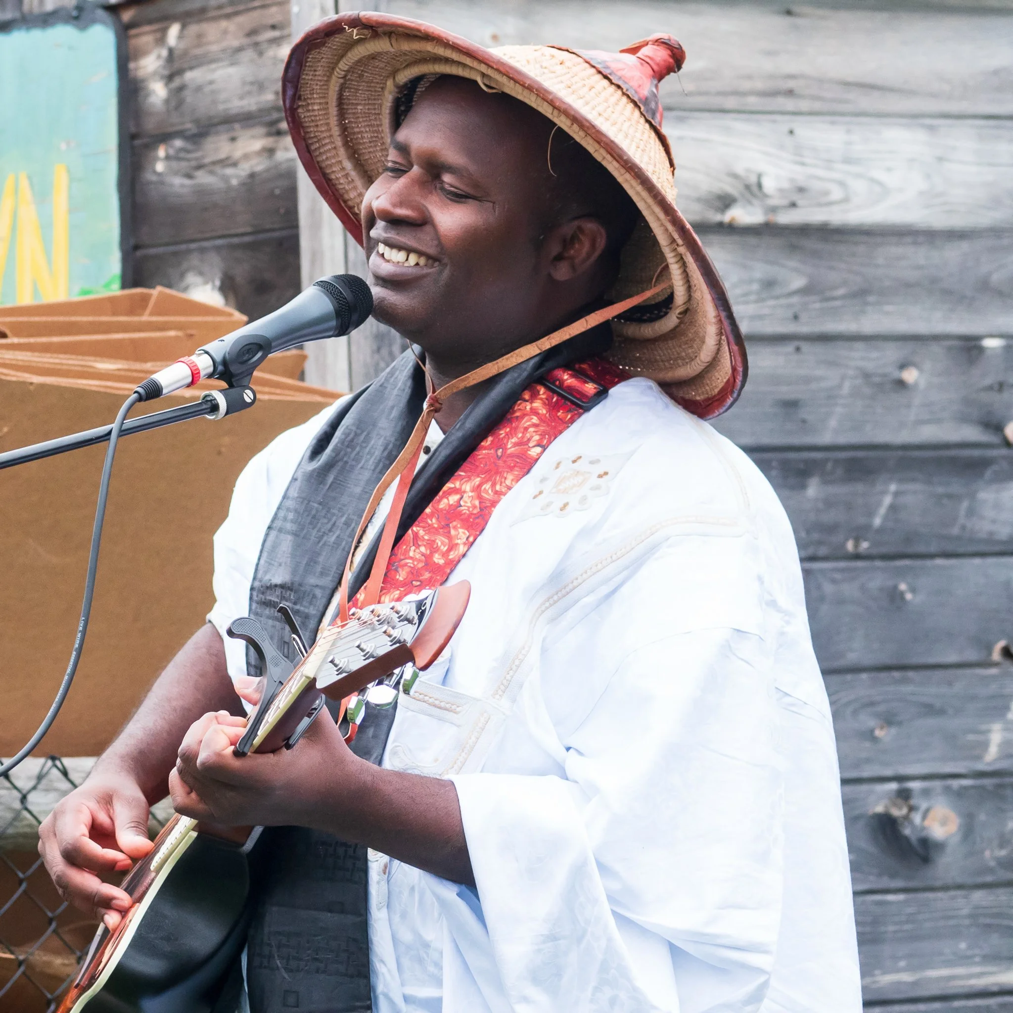 A man singing into a microphone while playing an acoustic guitar, wearing traditional attire and a large straw hat.