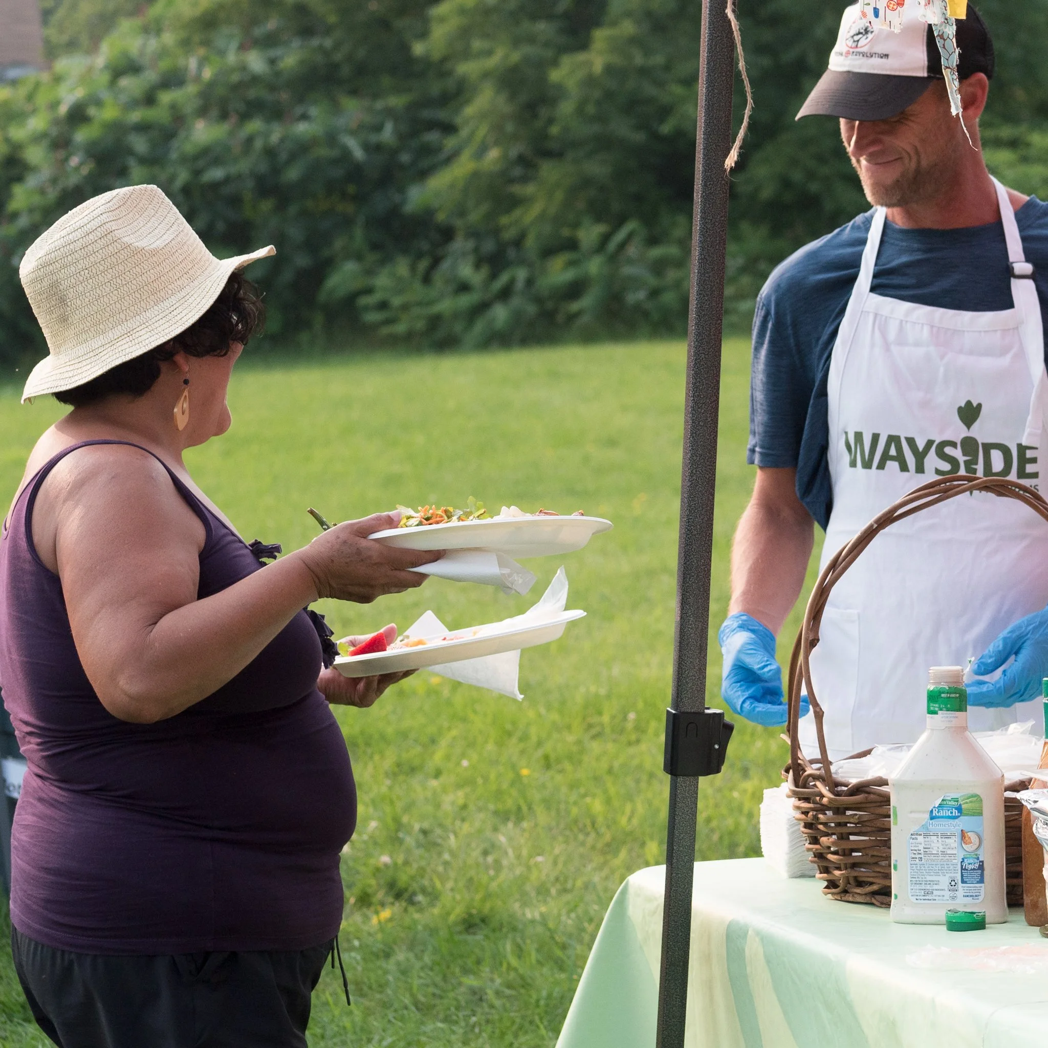 Woman wearing a sun hat and purple sleeveless top receives food trays from man in a white apron and blue gloves at an outdoor food stand in a grassy area.