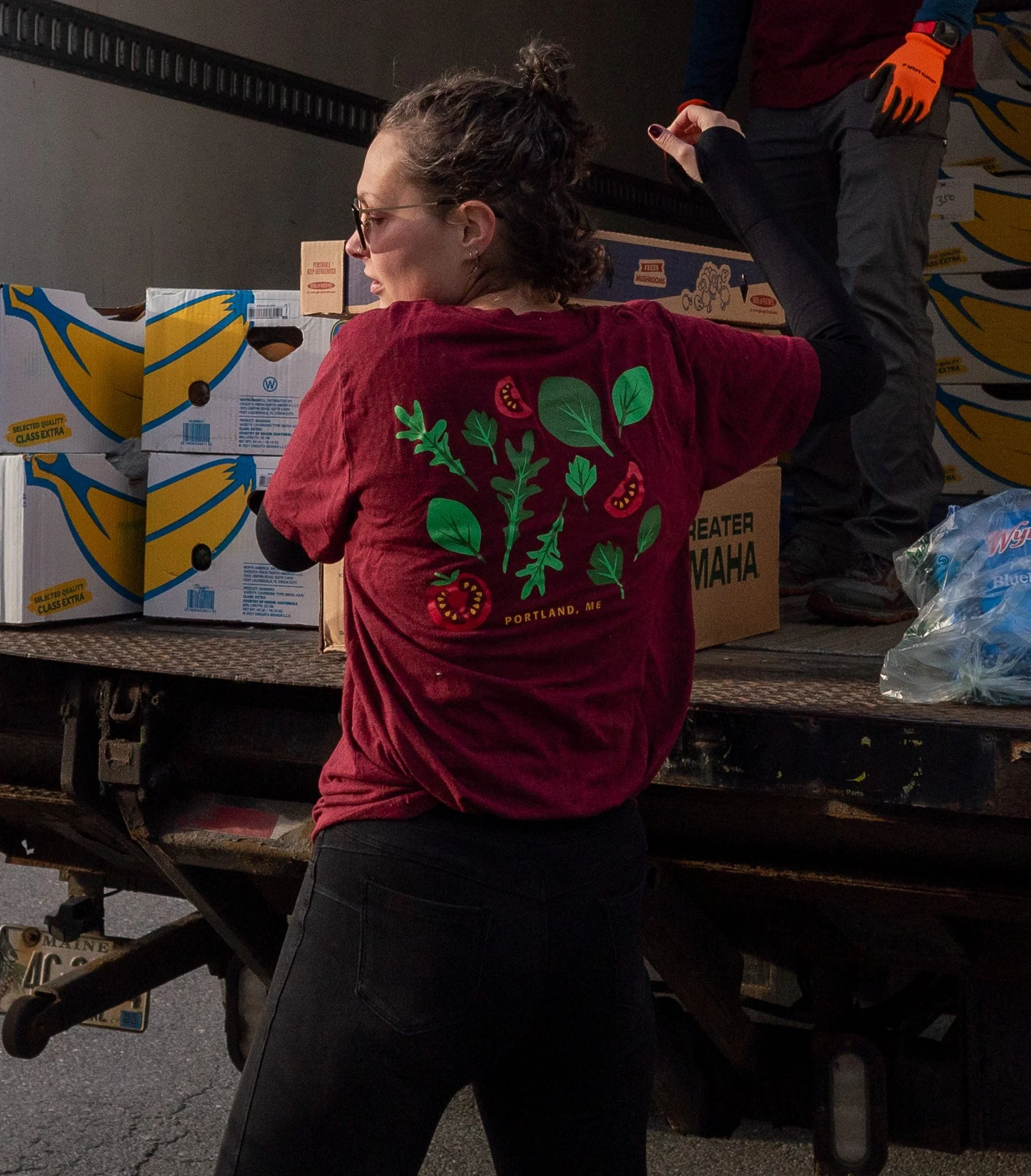 A woman with curly hair and glasses wearing a red t-shirt with a green leafy design and the words 'Portland, ME' is loading boxes on a truck. Another person, partially visible, is standing on the truck, wearing gray pants and orange gloves.