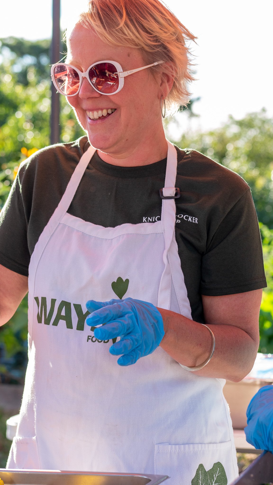 A woman with short blonde hair wearing sunglasses, a black shirt, and a white apron with 'WAY' and a green heart logo, is smiling and wearing blue gloves while preparing food outdoors.