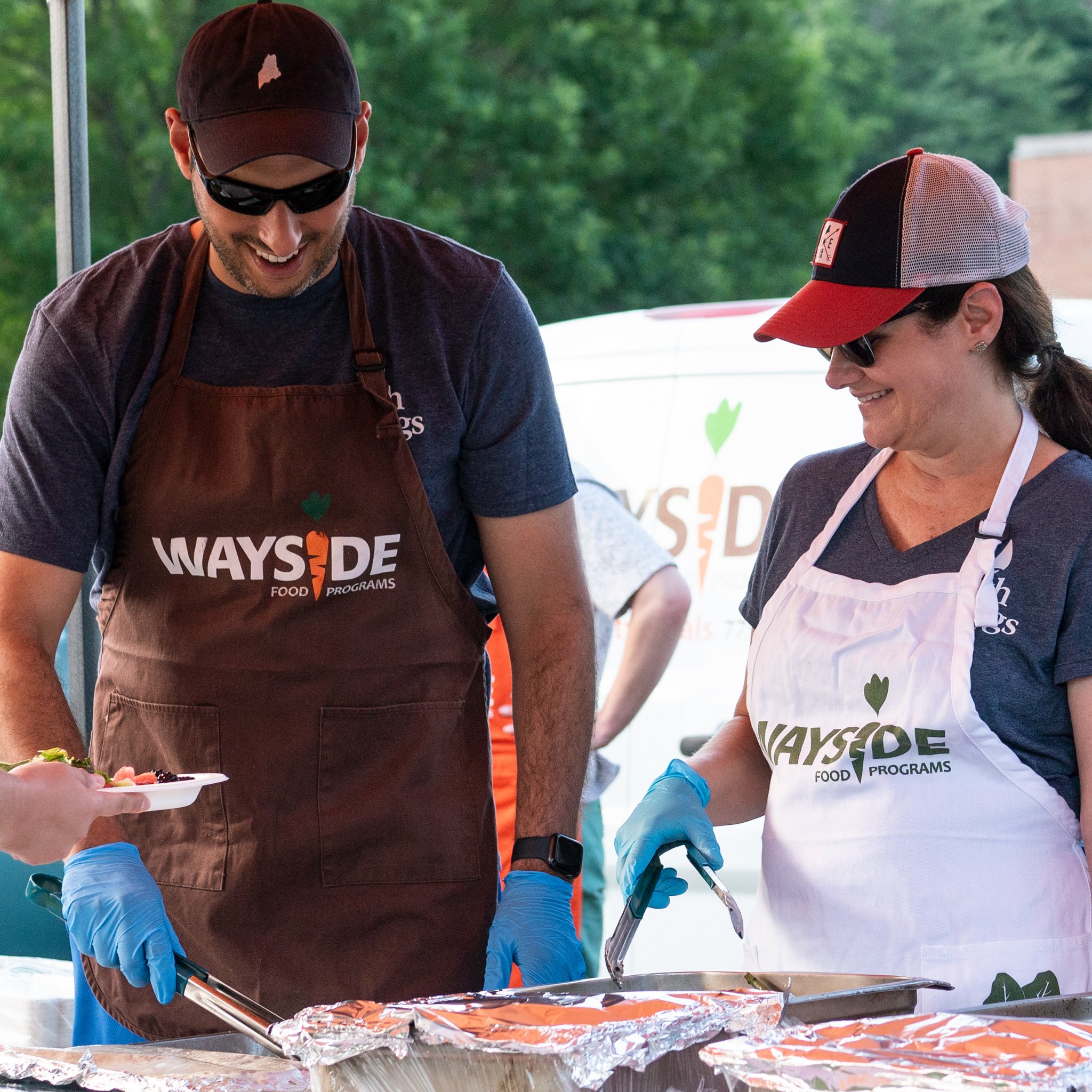 Two people serving food at a community event, wearing aprons with the "Wayside Food Programs" logo, outdoors with a white van in the background.