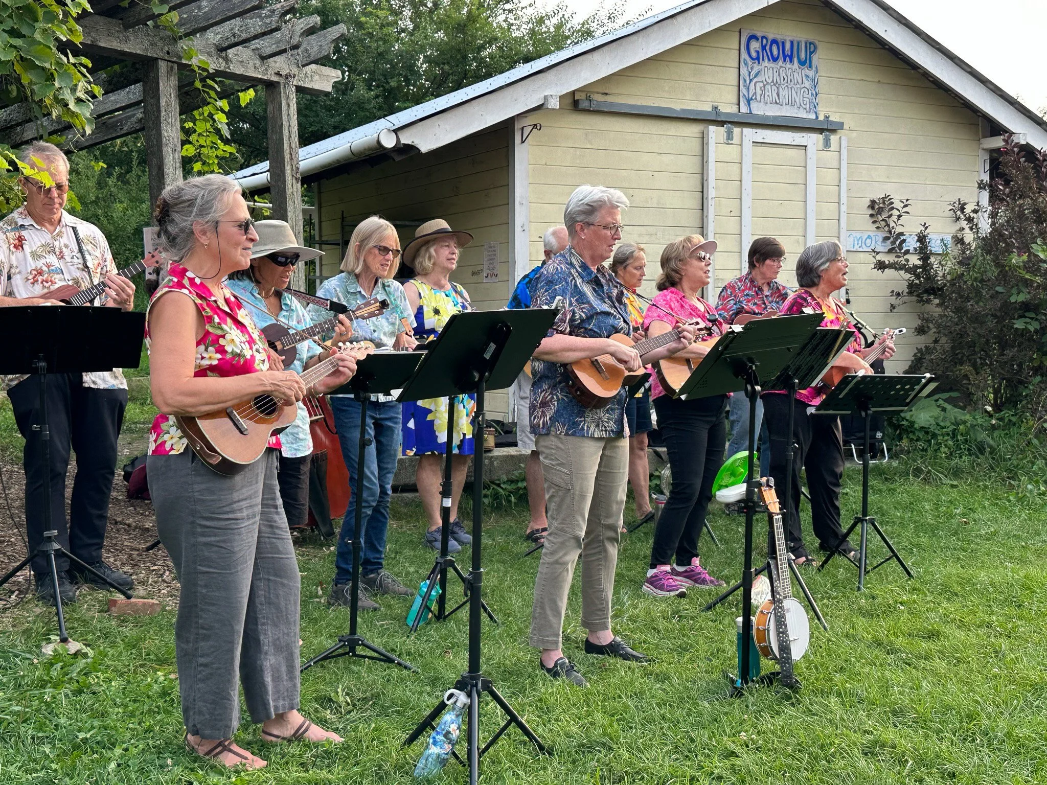 A group of older adults playing ukuleles outdoors in a yard, standing in front of a small building with a sign that says 'GROW UP', surrounded by greenery.