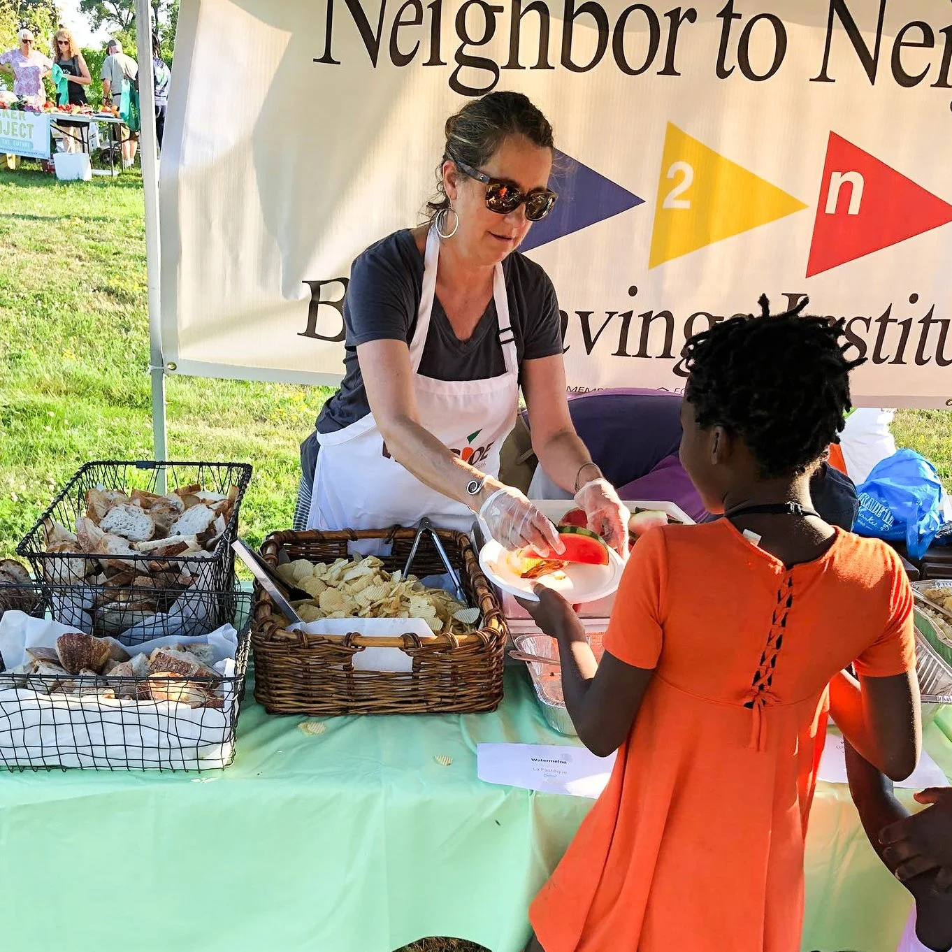 A woman serving food to a girl at an outdoor event, with a sign that reads "Neighbor to Neighbor" in the background.