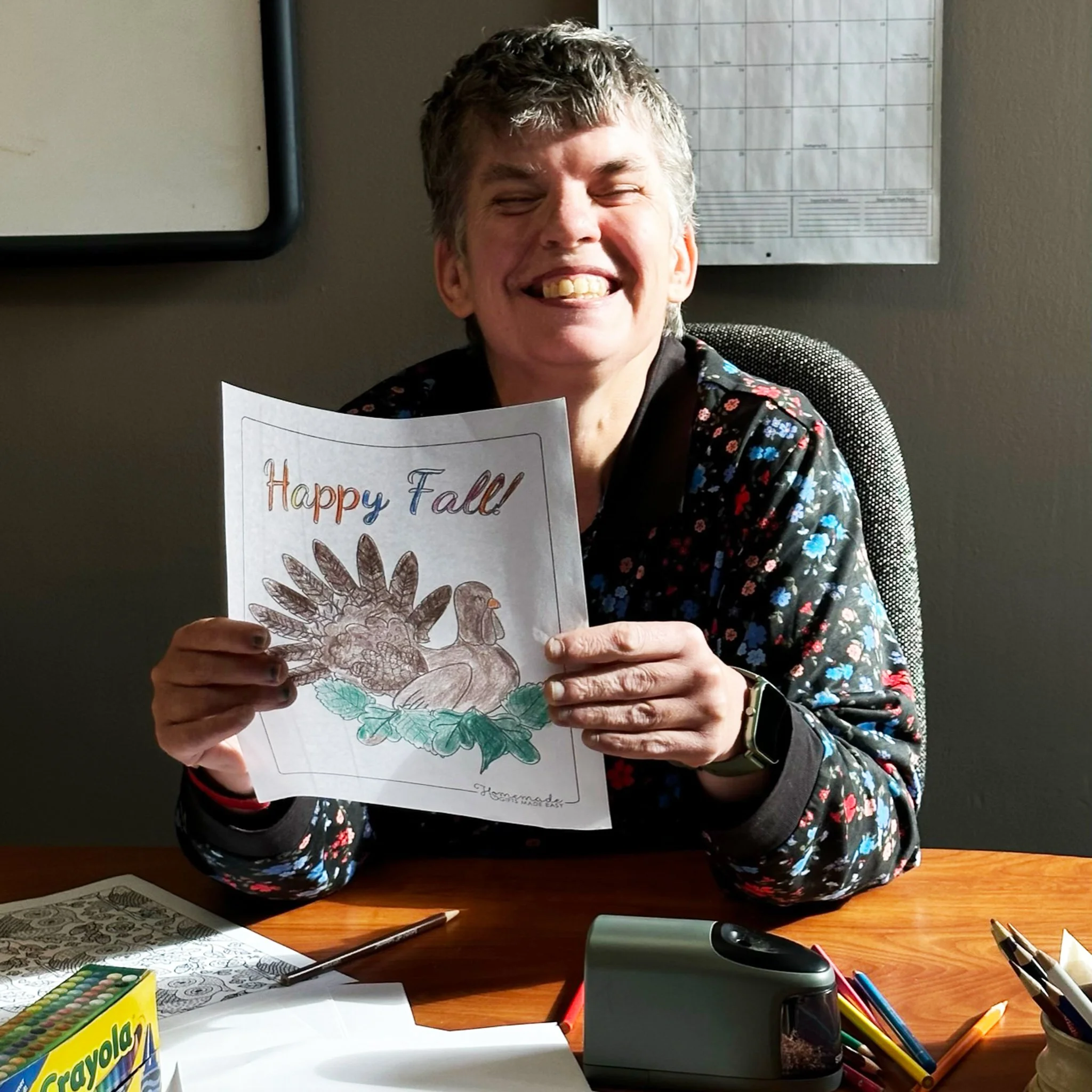 Woman smiling and holding a Thanksgiving card with a drawing of a turkey and fall leaves, sitting at a desk with coloring supplies.