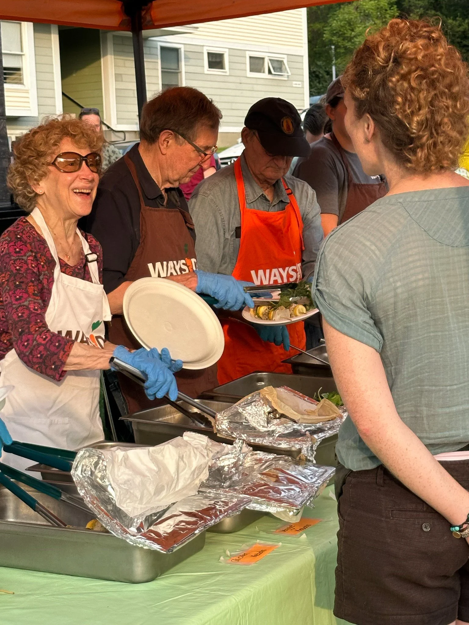 People serving and receiving food at a buffet table during an outdoor event, with some wearing aprons and gloves.