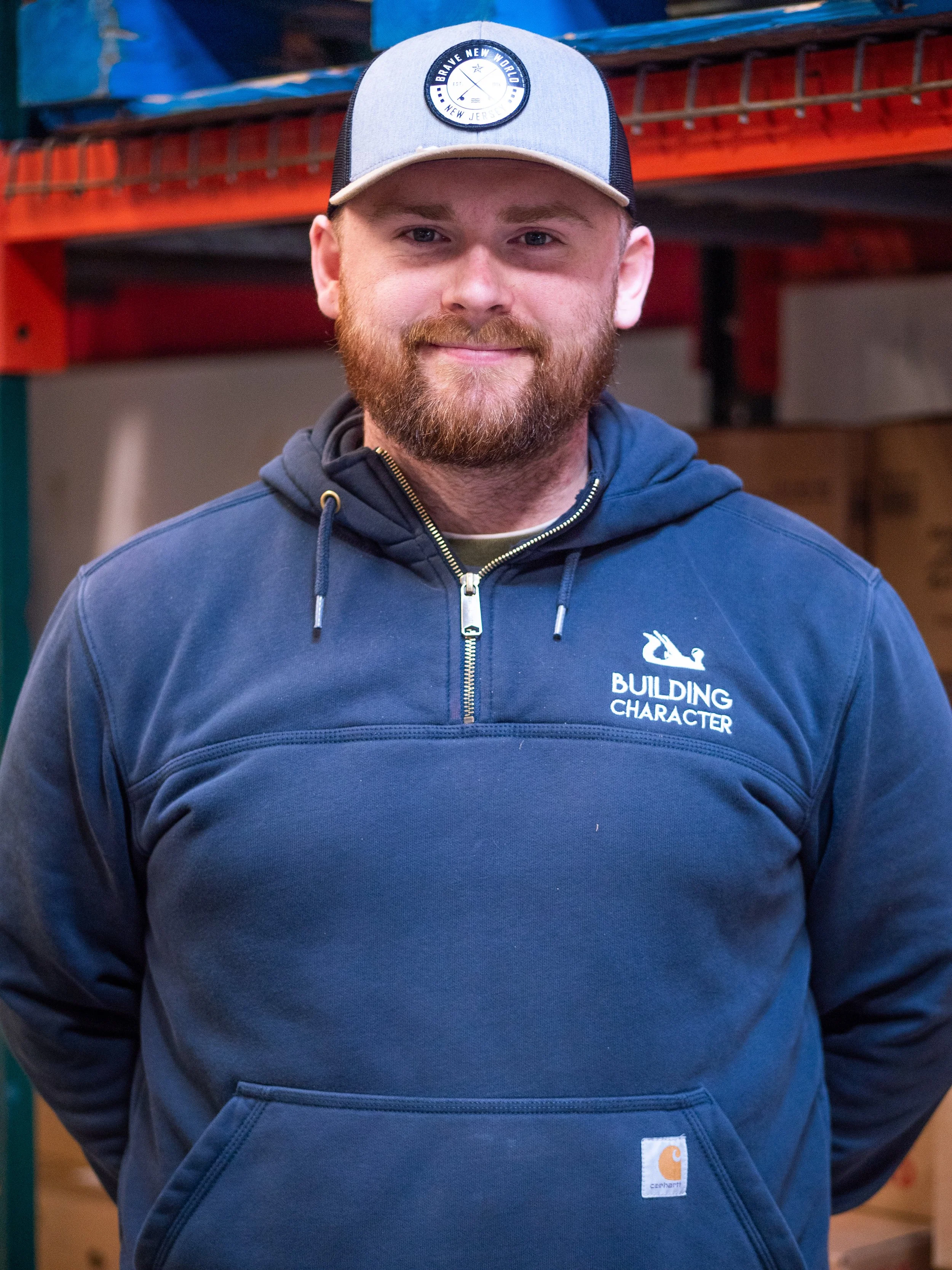 A young man with a beard and mustache wearing a blue hoodie and a gray and black baseball cap with a logo, standing indoors in front of a shelving unit.