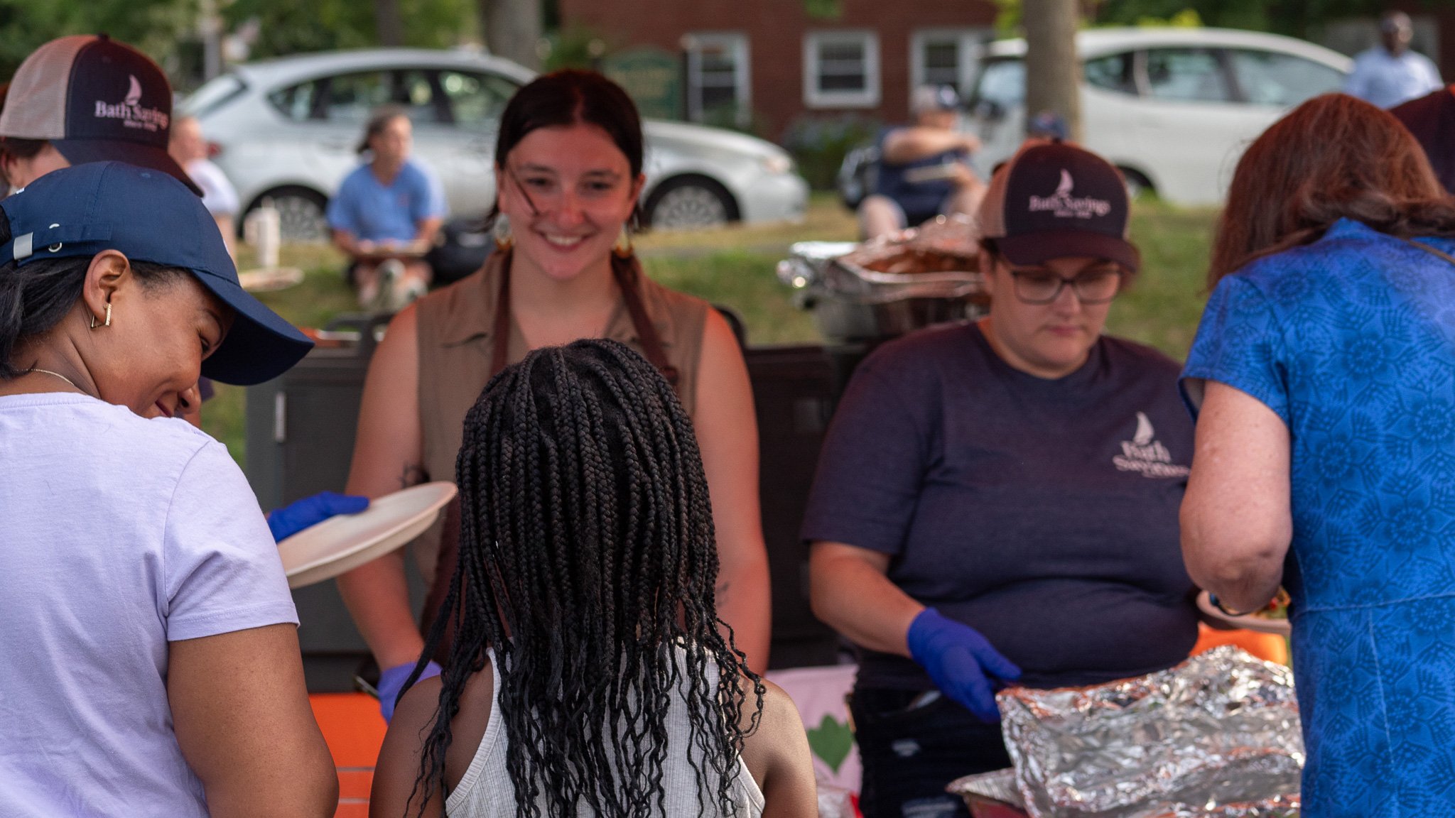 People standing in front of a food stand outdoors, preparing and serving food, with cars and trees in the background.