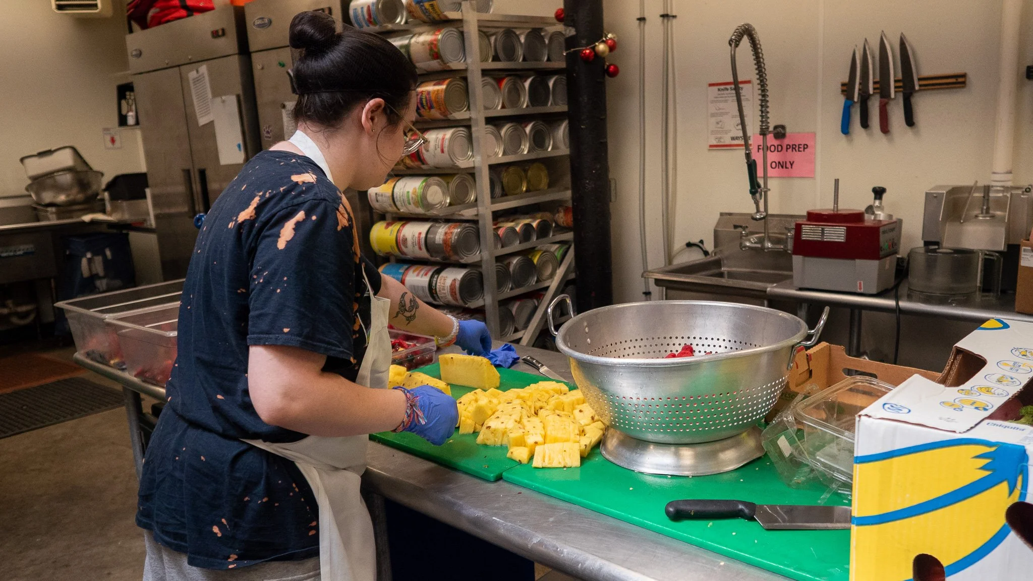 A woman with dark hair tied in a bun, wearing glasses, a navy blue shirt with paint splatters, a white apron, and blue gloves, is chopping pineapple on a green cutting board in a commercial kitchen.