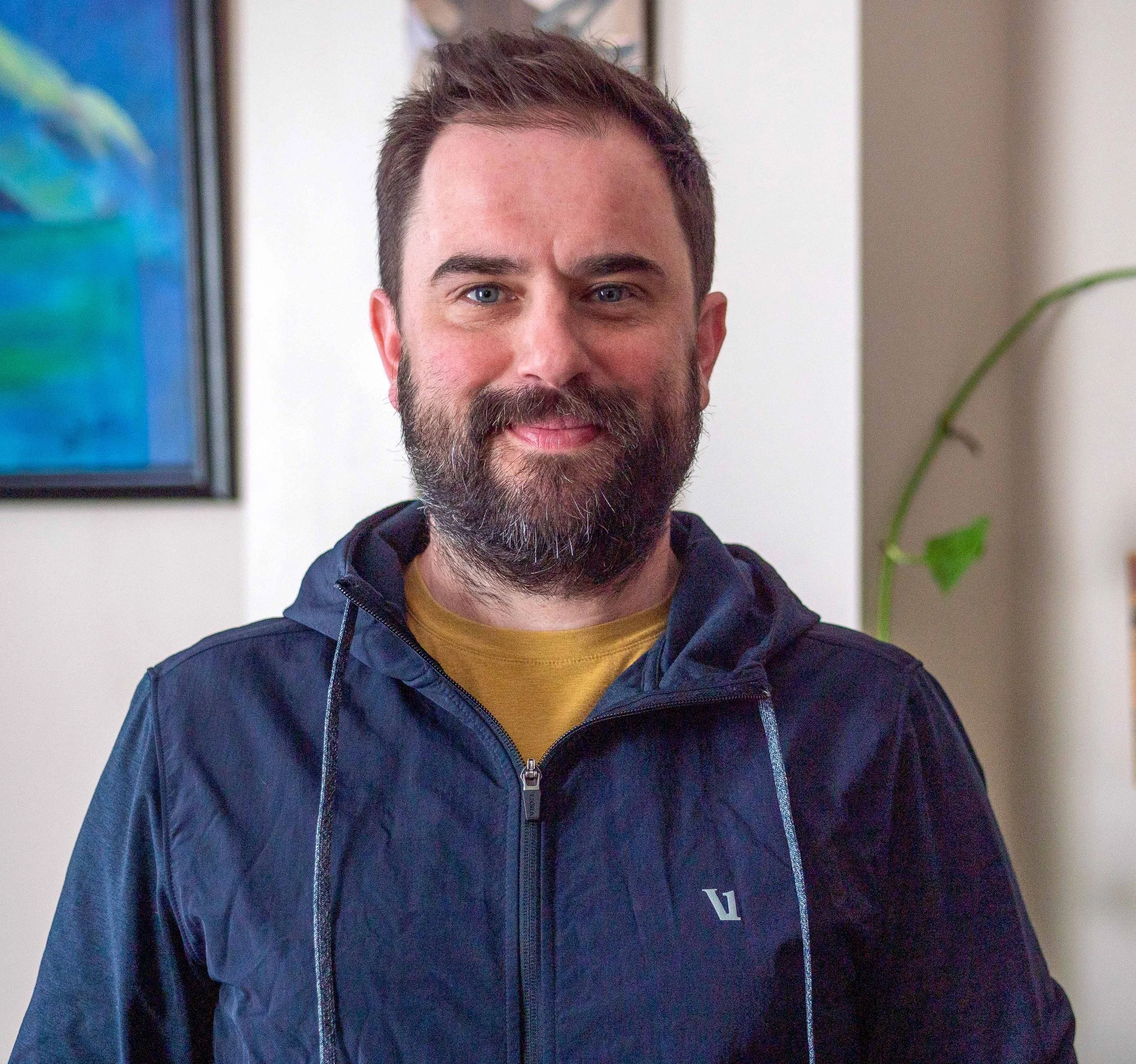 A smiling man with a beard and short brown hair, wearing a navy blue jacket with a yellow shirt underneath, standing indoors with a framed painting and a green plant in the background.