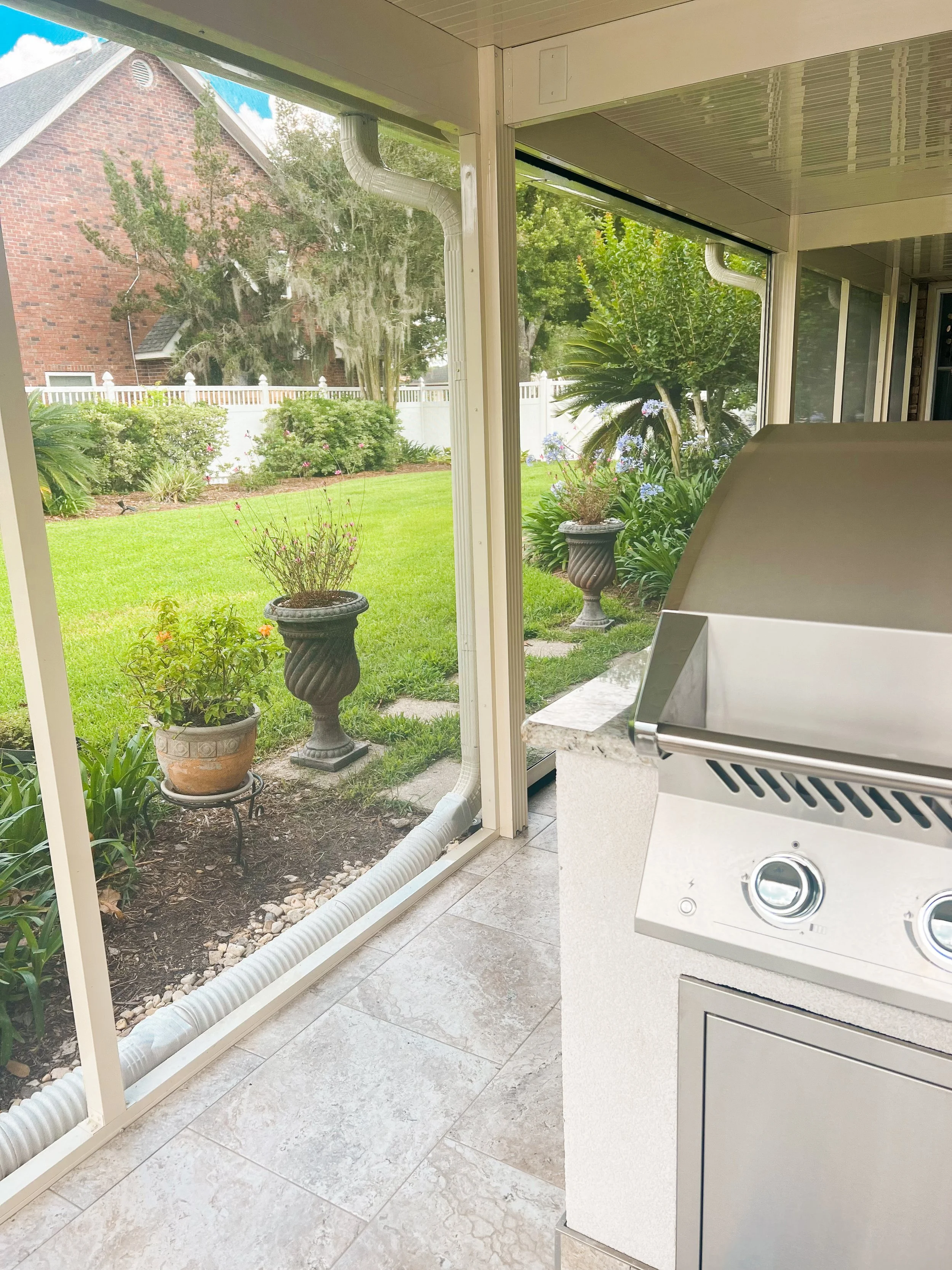 Outdoor patio with a stainless steel grill, tiled floor, and enclosed glass walls overlooking a garden with potted plants and a brick house in the background.
