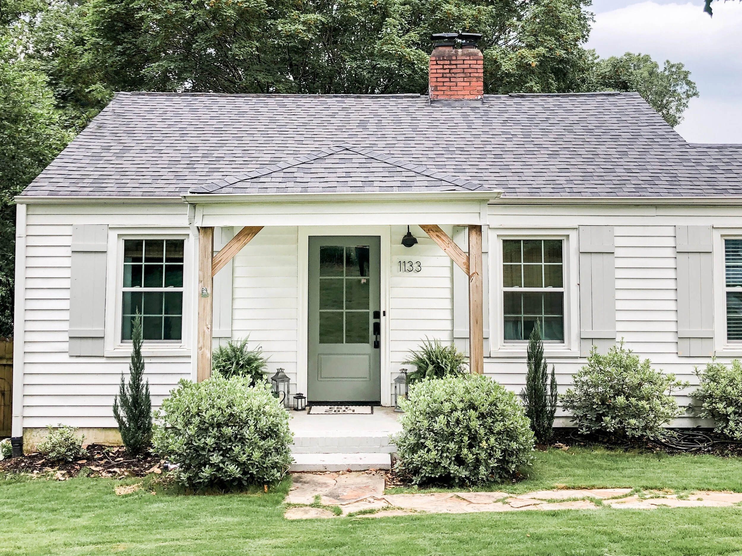 Small white house with gray shutters, a central front door, and a brick chimney. The yard has green grass and small bushes near the entrance.