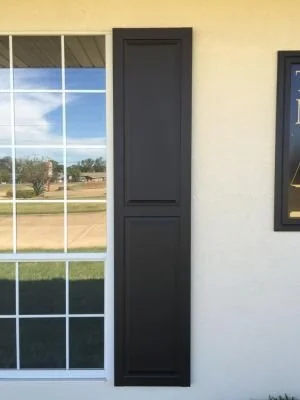 Black window shutter next to a large window with a grid pattern on a beige wall.