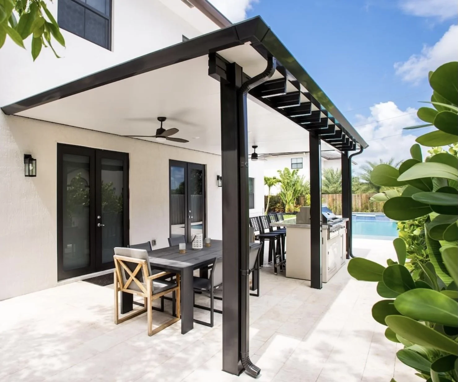 Modern backyard patio with outdoor dining table and chairs under a covered pergola, next to a barbecue grill and seating area. Features a pool in the background, surrounded by plants and trees.