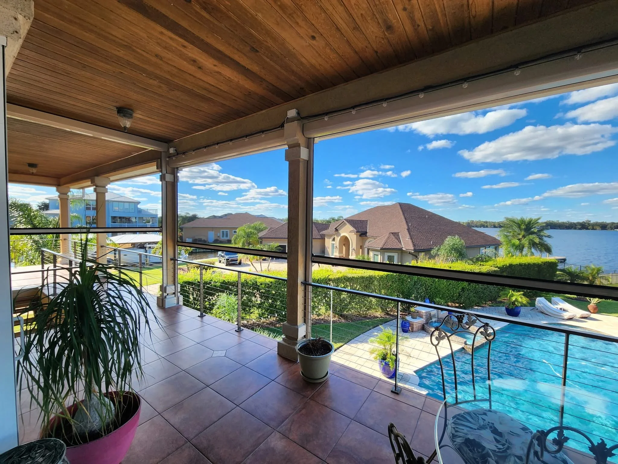 Outdoor patio with wooden ceiling, tiled floor, and railing overlooking a backyard with a pool. View includes a lake, surrounding houses, lush greenery, and a bright blue sky with clouds.
