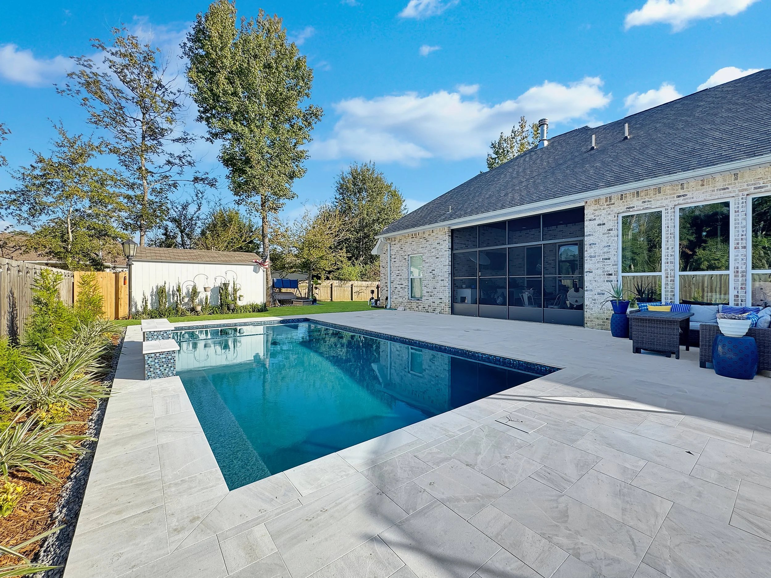 A backyard scene featuring a swimming pool surrounded by a stone-tiled patio. The patio is adjacent to a modern house with large windows and outdoor furniture. There are trees and plants along the fence, adding greenery to the space. The sky is clear and blue.