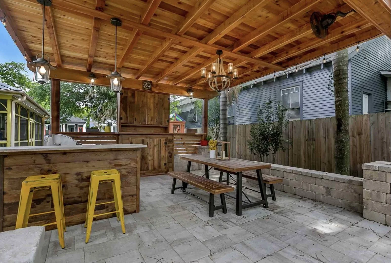 Outdoor patio with wooden pergola, three industrial pendant lights, picnic table set, yellow bar stools, and kitchen counter. Surrounded by a wooden fence with lush greenery.