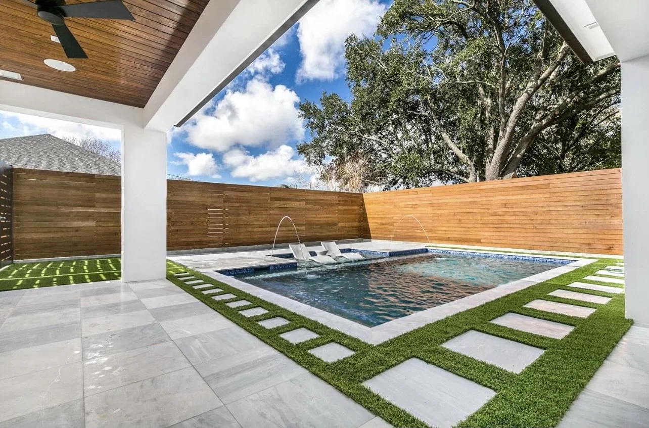 Modern backyard with a swimming pool, lounge chairs, fountain features, and surrounding wooden fence. The area includes a covered patio with a ceiling fan and a tiled floor with grass accents.