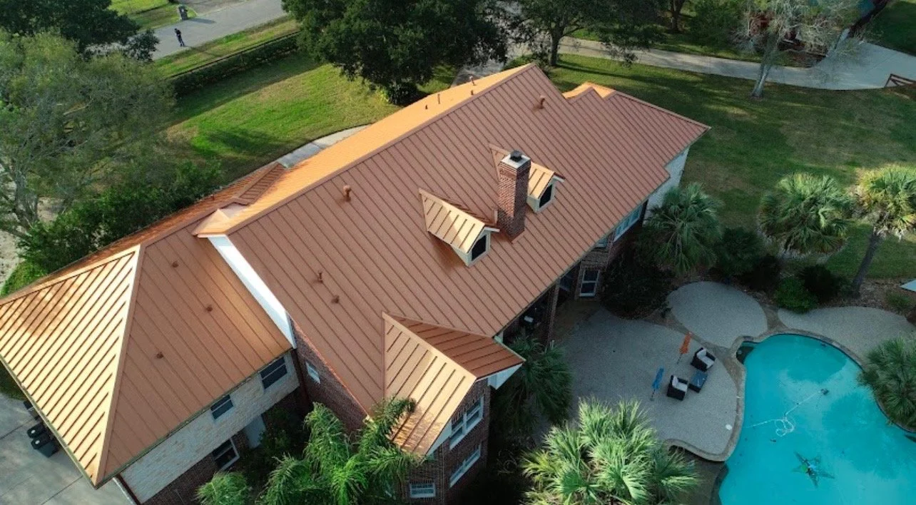 Aerial view of a house with a copper-colored metal roof, brick exterior, and a swimming pool in the backyard surrounded by palm trees and greenery.