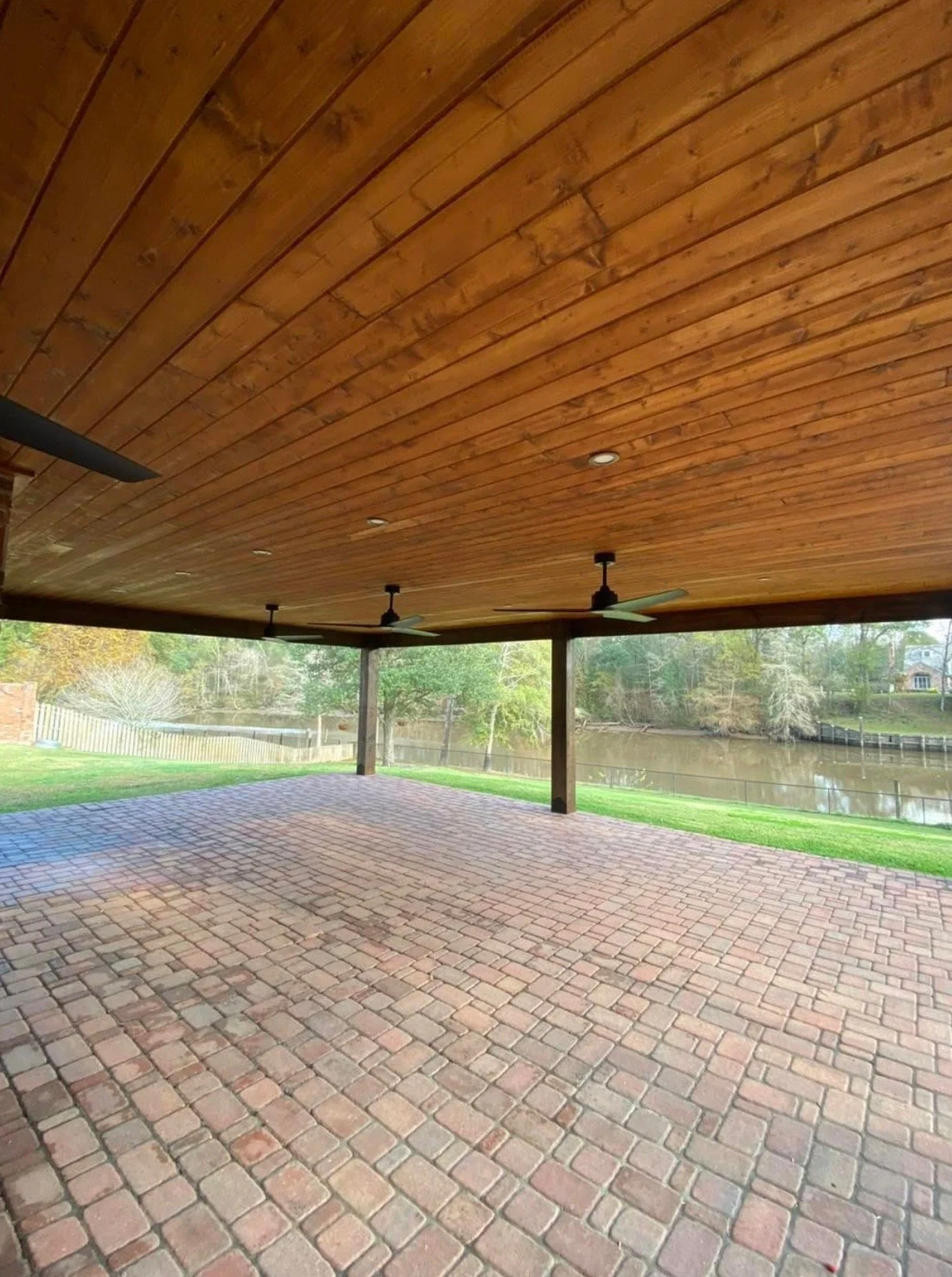Covered patio with brick flooring, wooden ceiling, ceiling fans, overlooking a grassy area and a river.