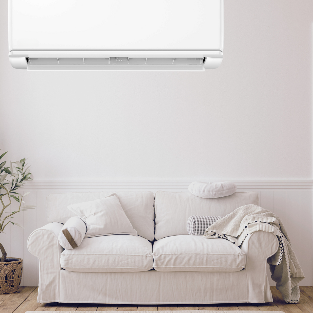 Witte bank met kussens en een deken in een woonkamer met houten vloer, witte muur en een groene plant in een mand, airconditioner boven de bank.