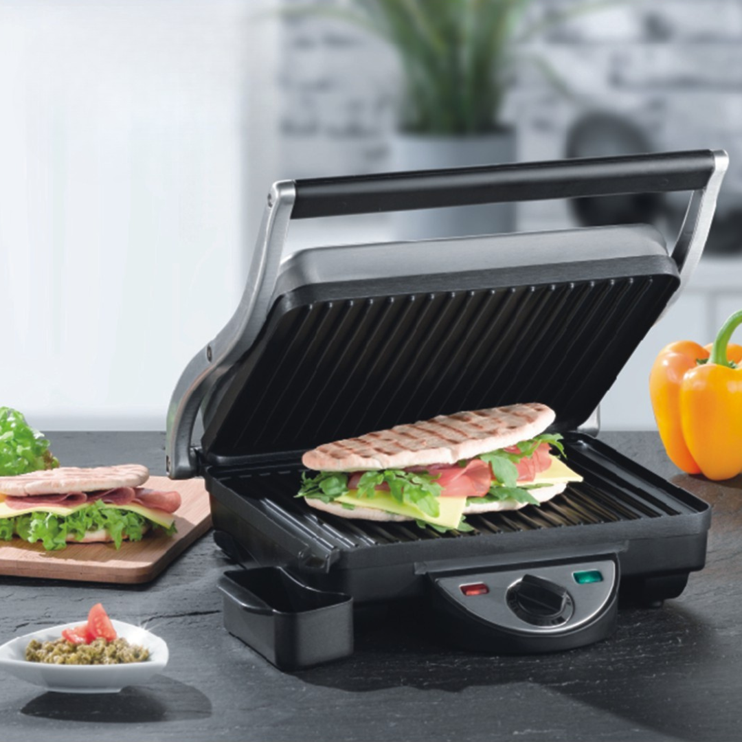 Een contactgrill met een gegrild broodje met ham, salami, kaas en sla, op een keukenblad, met, daarnaast, een bord met saus en bijgerechten, en paprika's op de achtergrond.