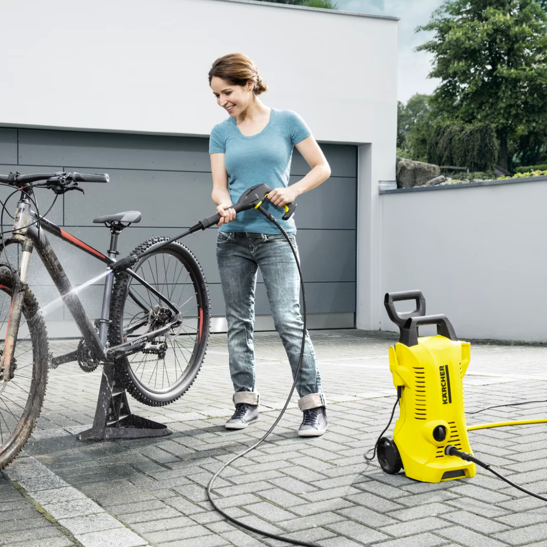 Een vrouw wast haar mountainbike met een hogedrukreiniger buiten op een bestraatte oprit.