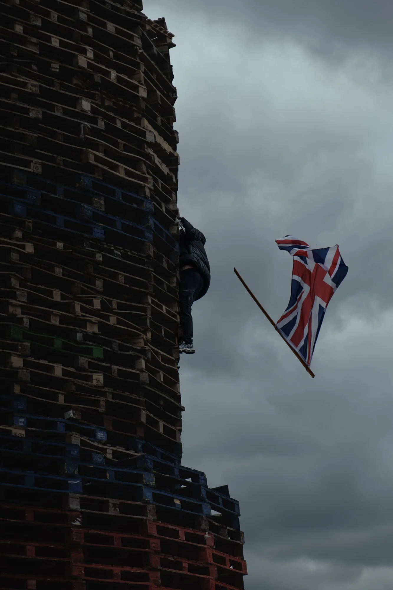Union Jack flag falls from a pyre as a youth climbs up, at Loyalist Sandy Row, Belfast. 2024