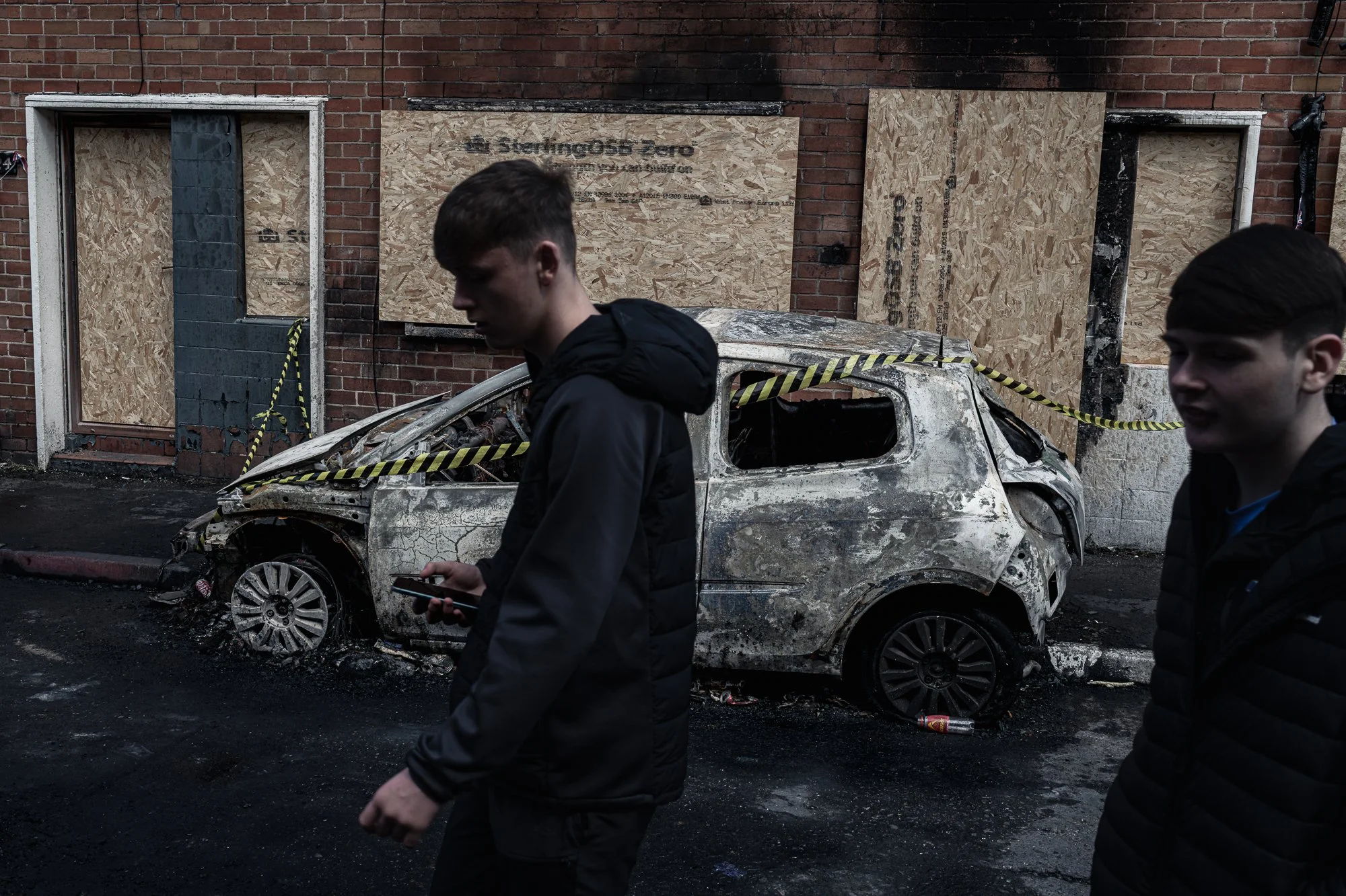 Teenagers walk past a burned vehicle in Newtownards Road after a night of rioting. Belfast. 2026