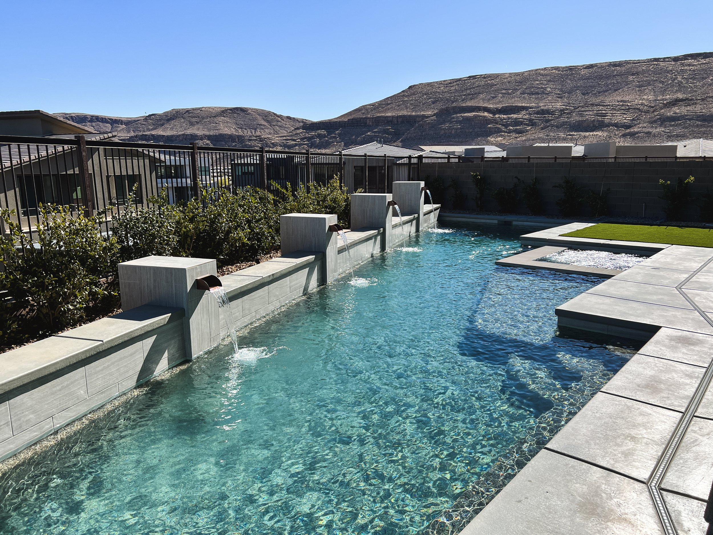 View of a Las Vegas backyard swimming pool with waterfalls, a gray stone deck, and a black metal fence. In the background, there are neighboring houses and a mountain range under a clear blue sky.
