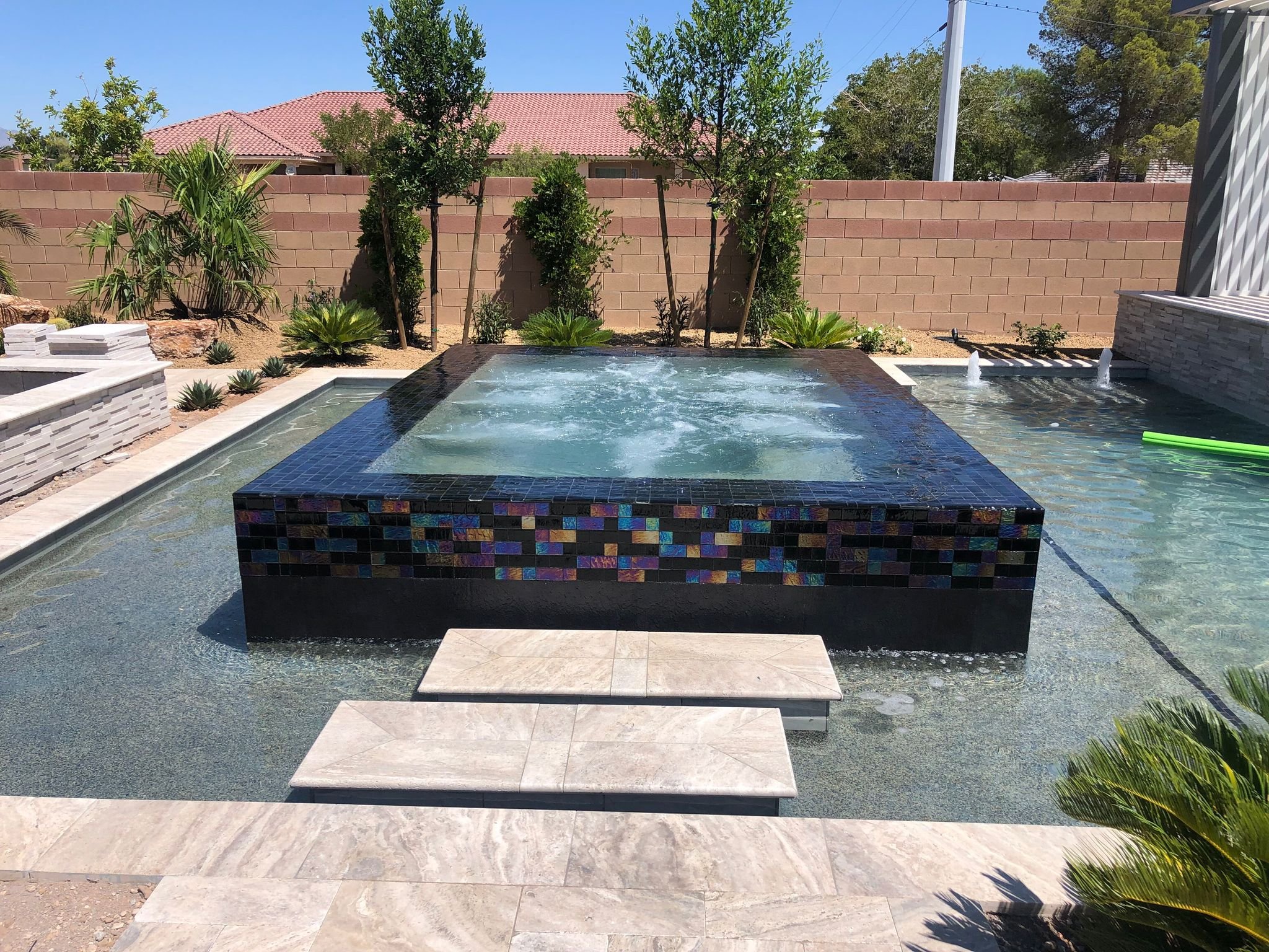 A Las Vegas backyard with a small pool or hot tub, a waterfall feature, tile accents, and surrounding greenery including trees and plants, enclosed by a brick wall.
