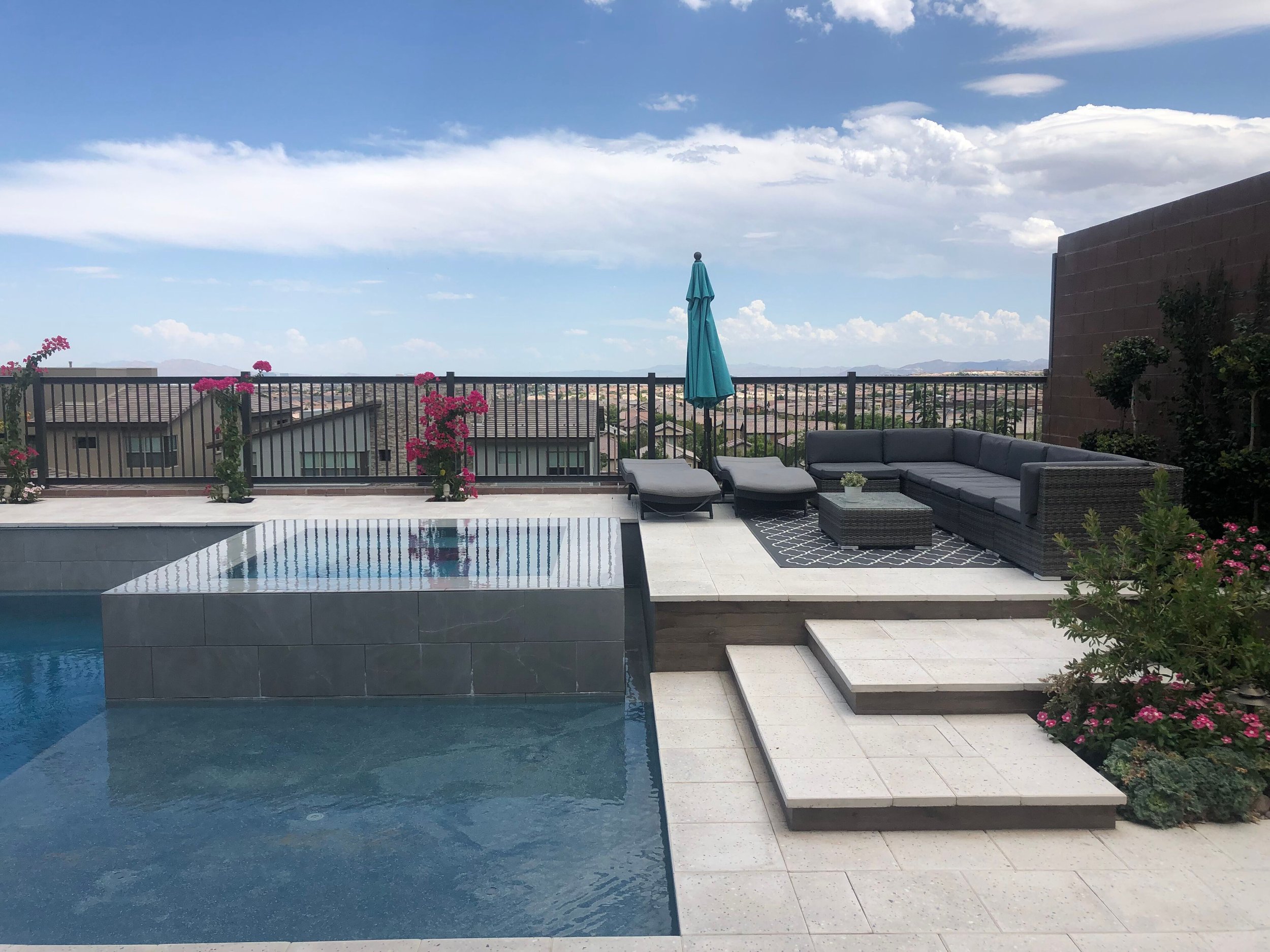 A Las Vegas outdoor rooftop patio with a swimming pool, lounge chairs, a patio sofa, pink flowering plants, a closed blue umbrella, and scenic mountain views under a partly cloudy sky.