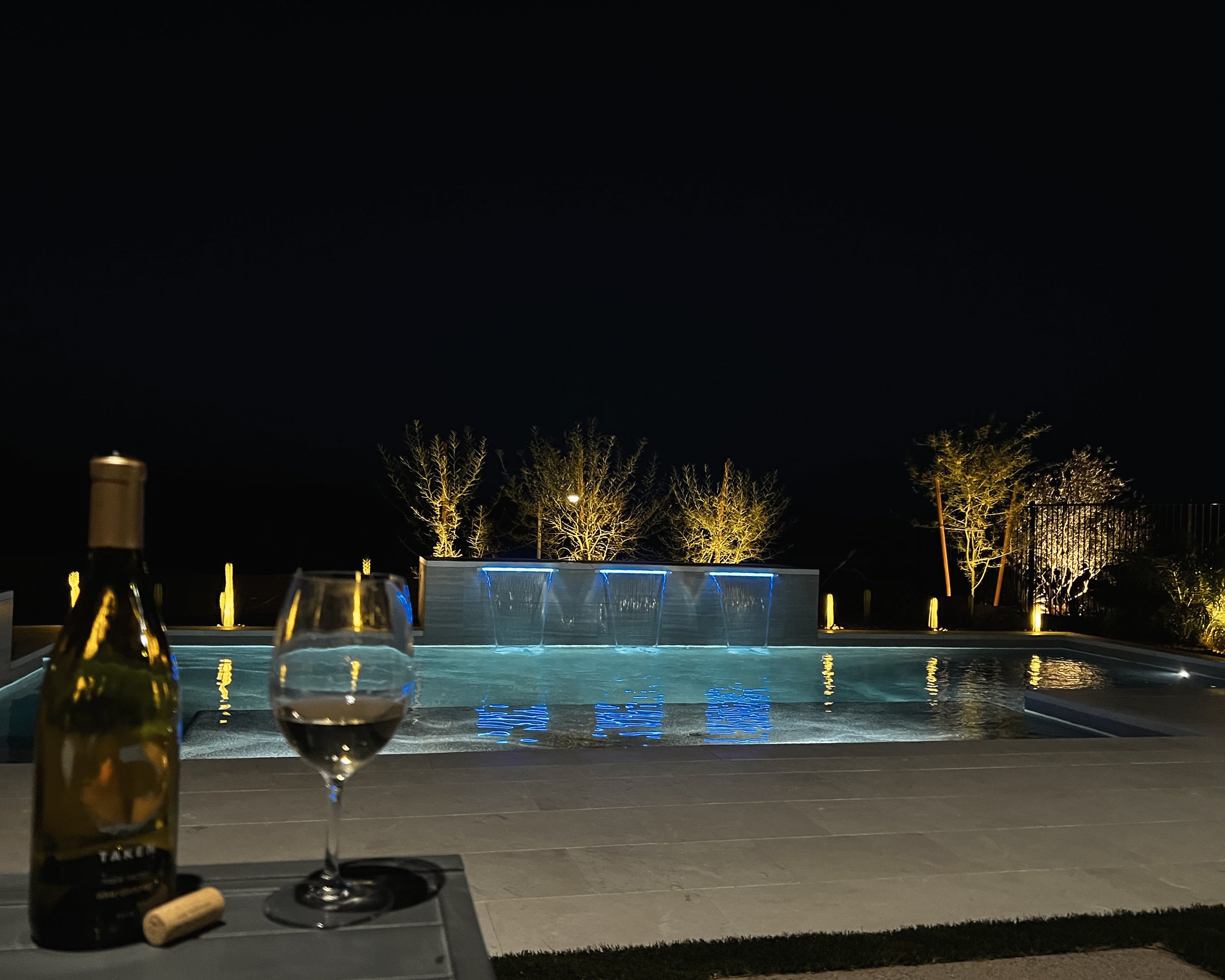 Nighttime view of a backyard with a swimming pool illuminated with blue lights, surrounded by trees and outdoor lighting. In the foreground, a table with two glasses of red wine and bottles, including a bottle of sparkling wine, and a cork. The scene