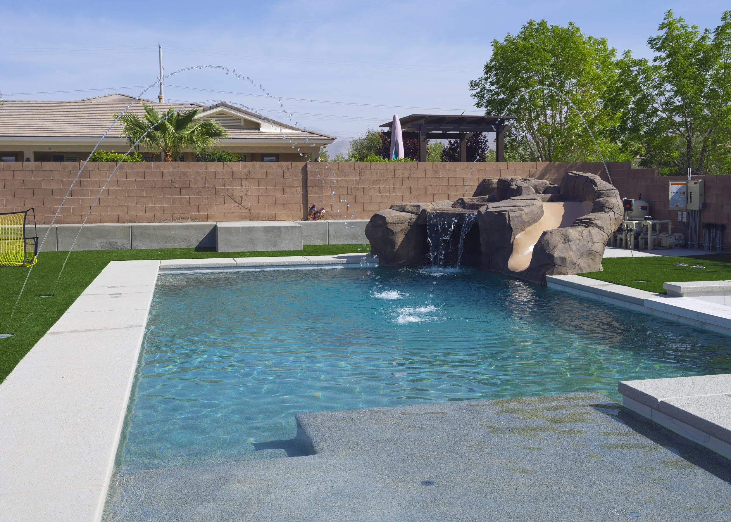 A swimming pool with a rock waterfall feature, surrounded by a concrete deck and green artificial grass, with a brick wall, trees, and a house in the background under a clear blue sky.
