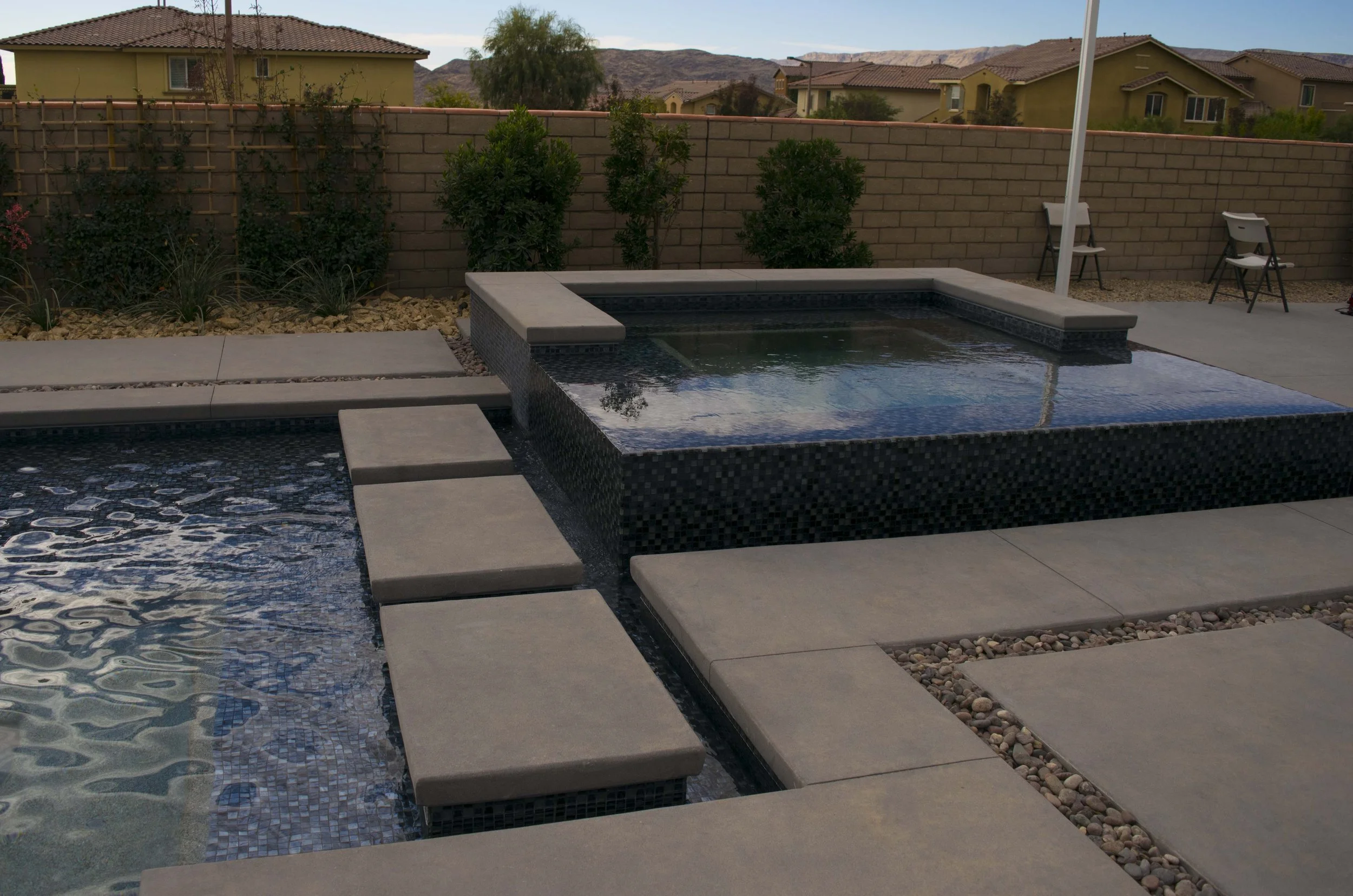 Backyard scene with a swimming pool featuring dark mosaic tiles, surrounded by a concrete deck. There are two white chairs with black legs and a white umbrella against a brick wall. Behind the wall, residential houses and mountains are visible in the