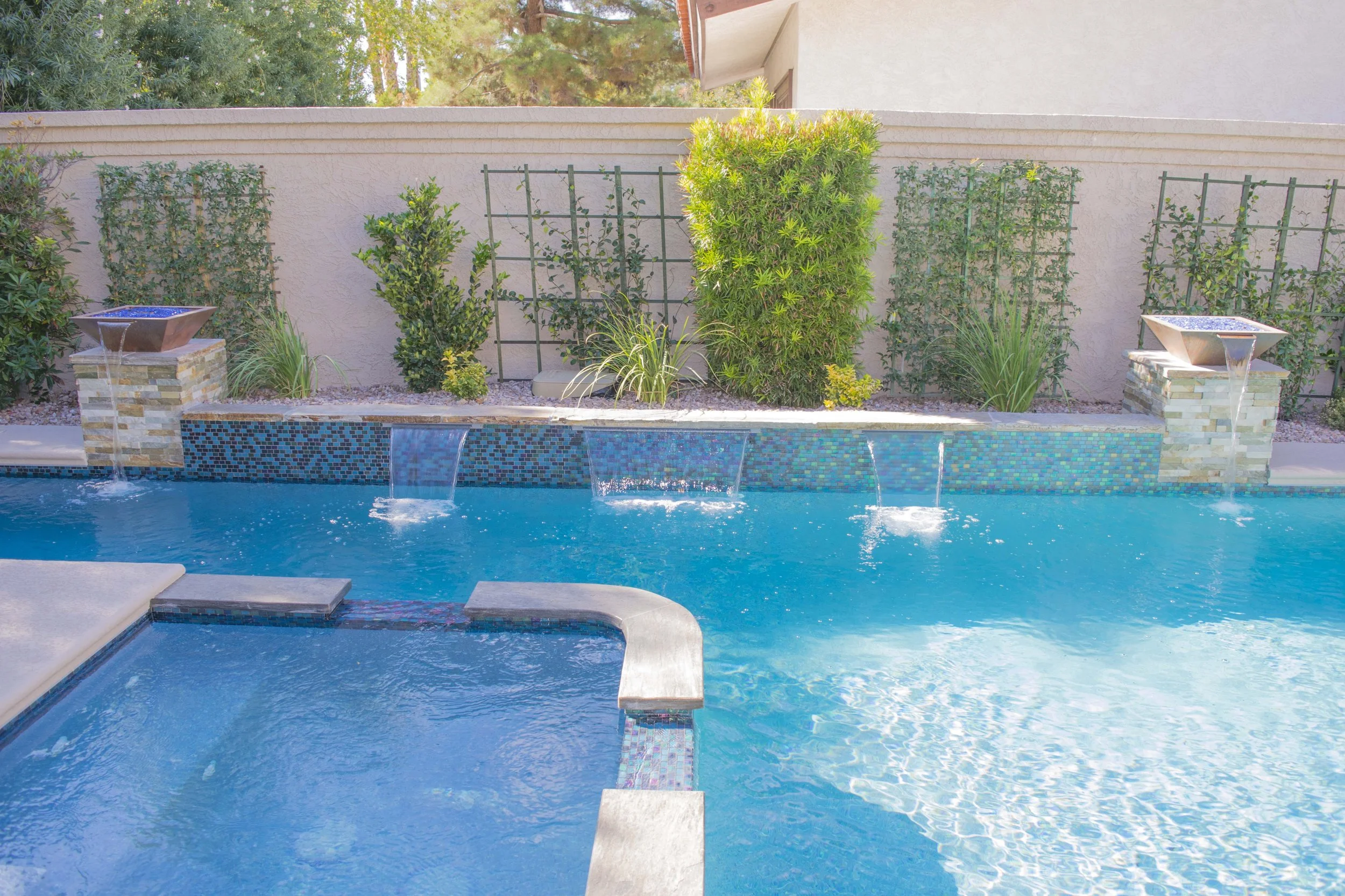 A Las Vegas backyard swimming pool with water features, surrounded by landscaping including bushes, trees, and trellises, with a beige stucco wall and house in the background.