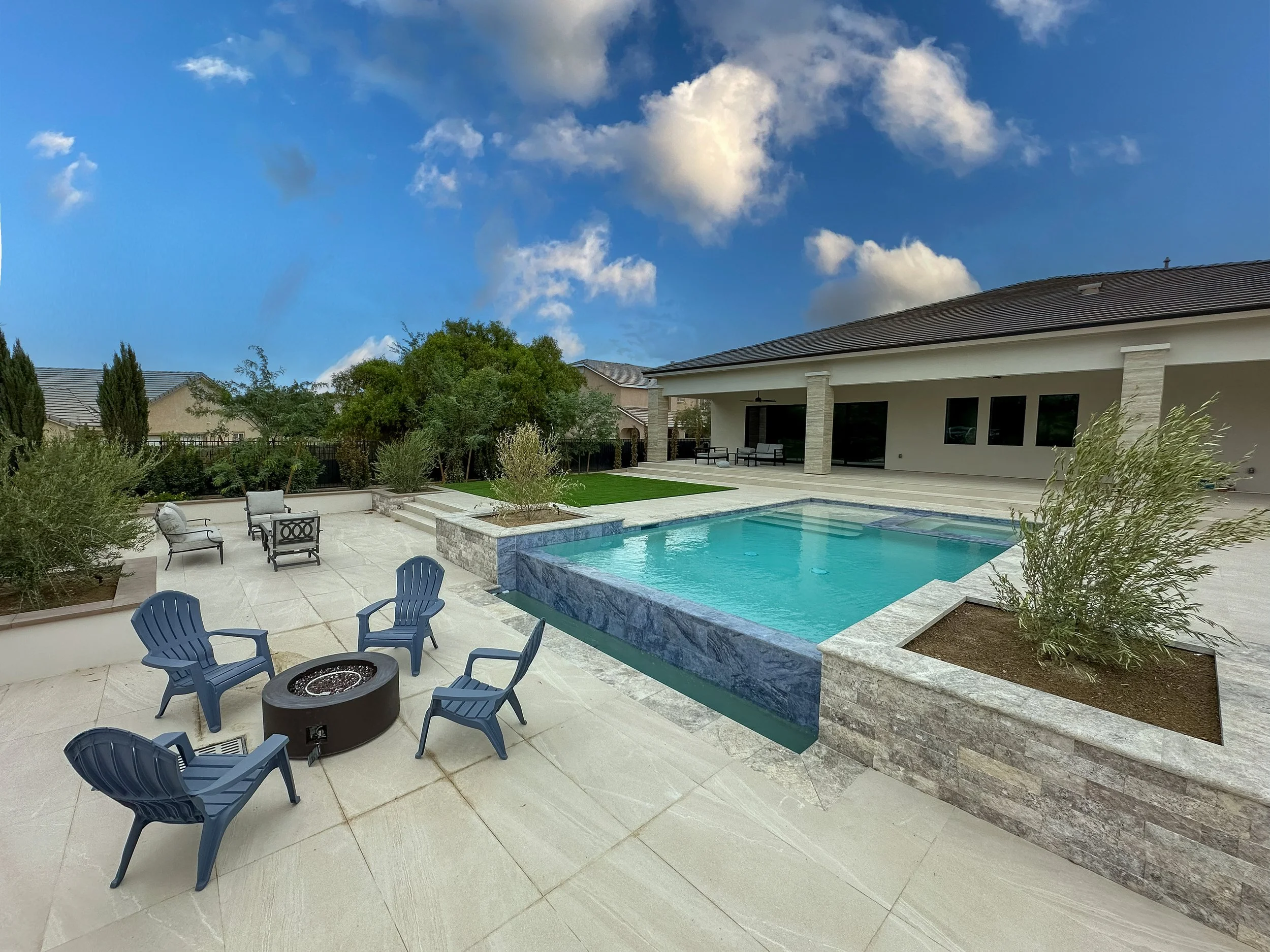 Las Vegas backyard with swimming pool, outdoor seating area with blue chairs, trees, and a house with a patio under a partly cloudy sky.