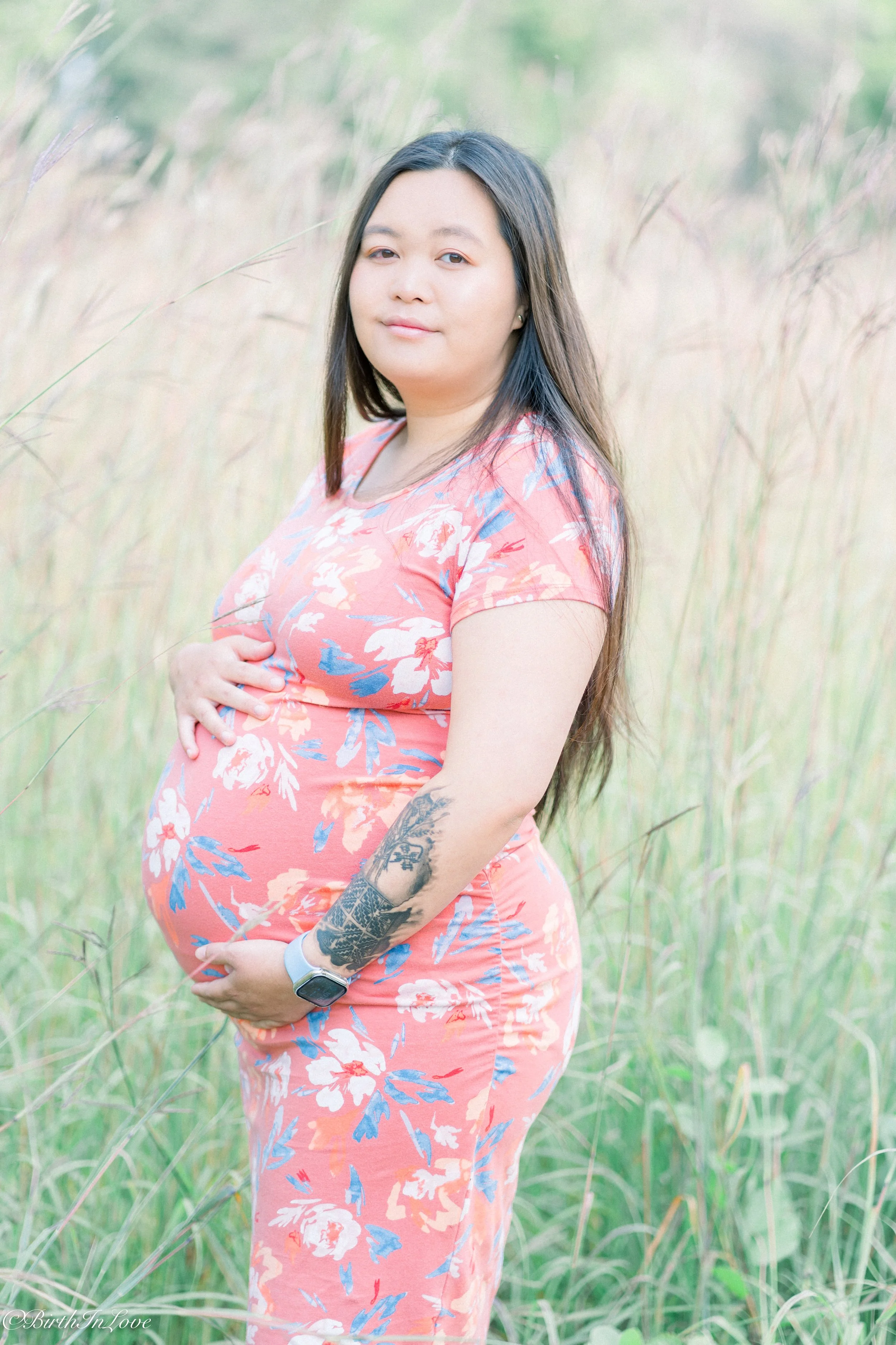 A pregnant woman with long dark hair standing in a grassy field, wearing a floral pink dress, with one hand on her belly and the other near her chest, looking at the camera with a subtle smile.