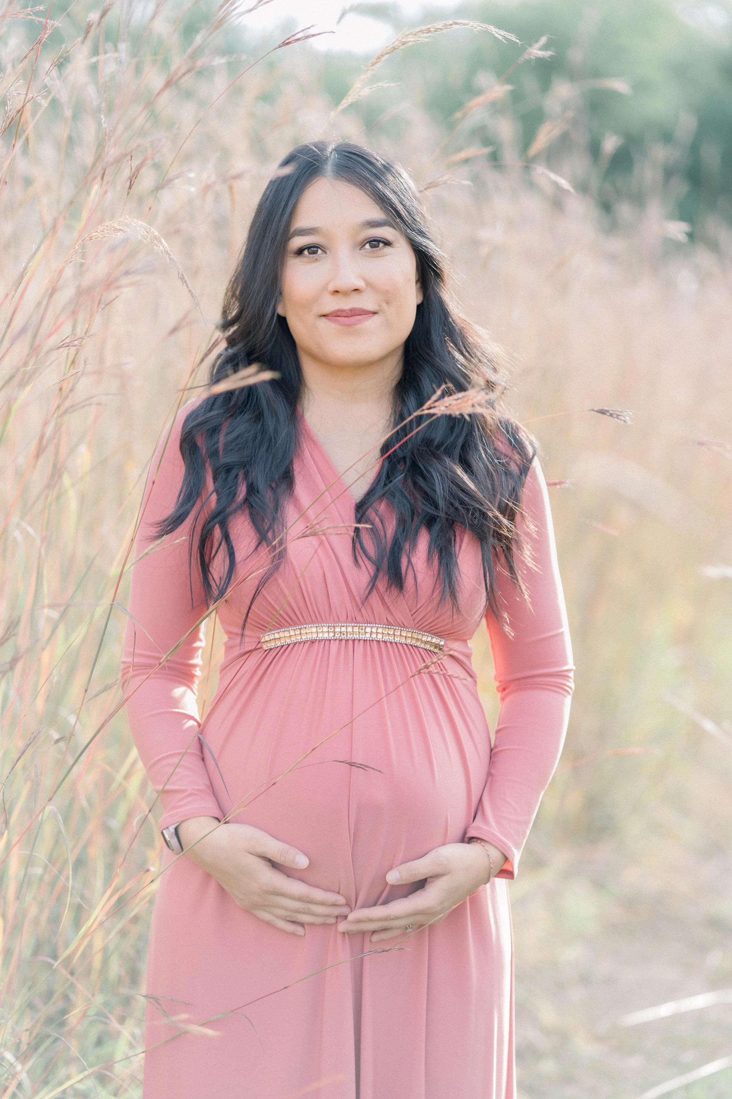 A pregnant woman with long dark hair in a pink dress standing in a field of tall grass.